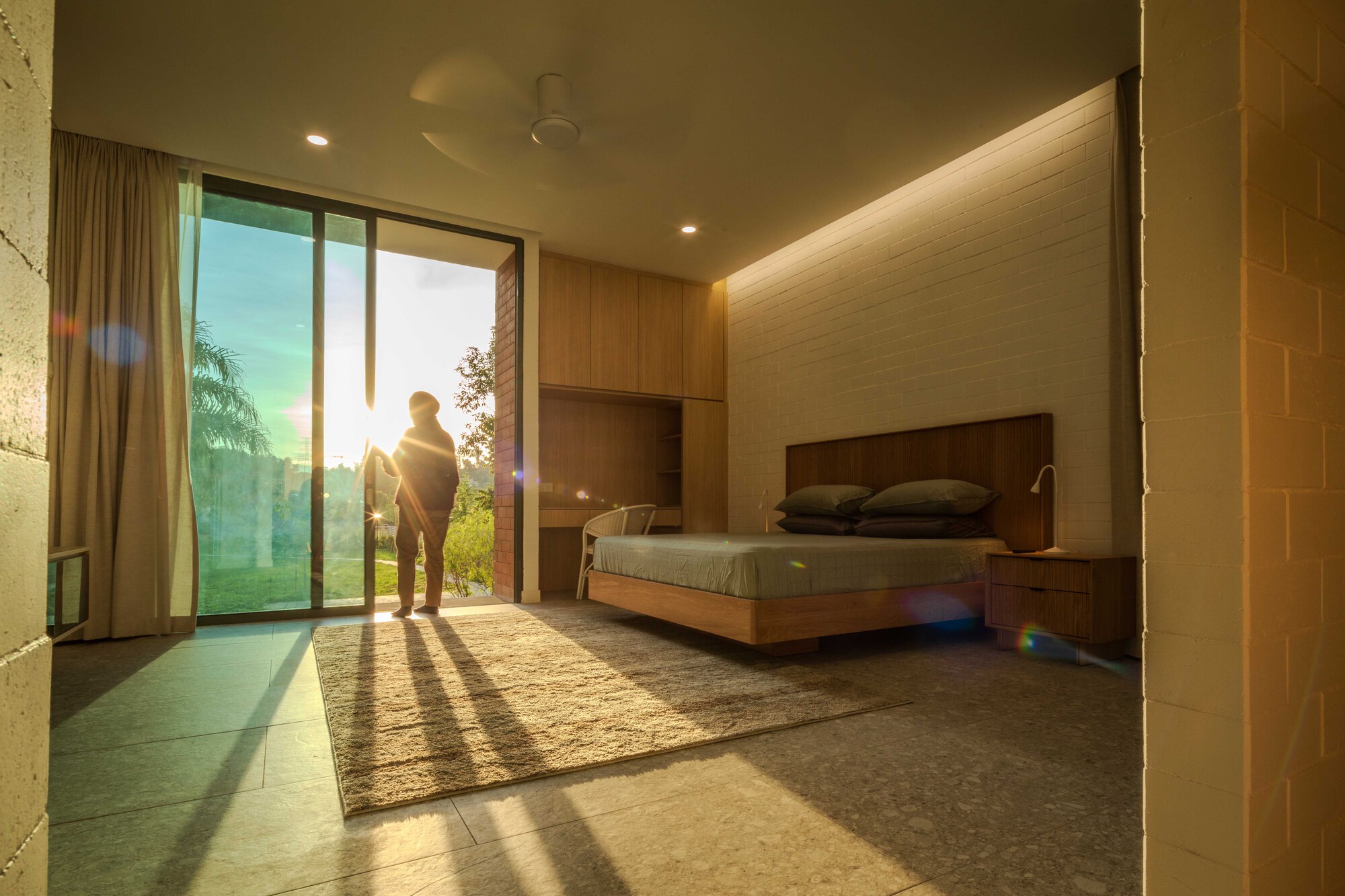 Bedroom interior with wood-paneled walls and full-height glazing framing two figures in golden sunset light