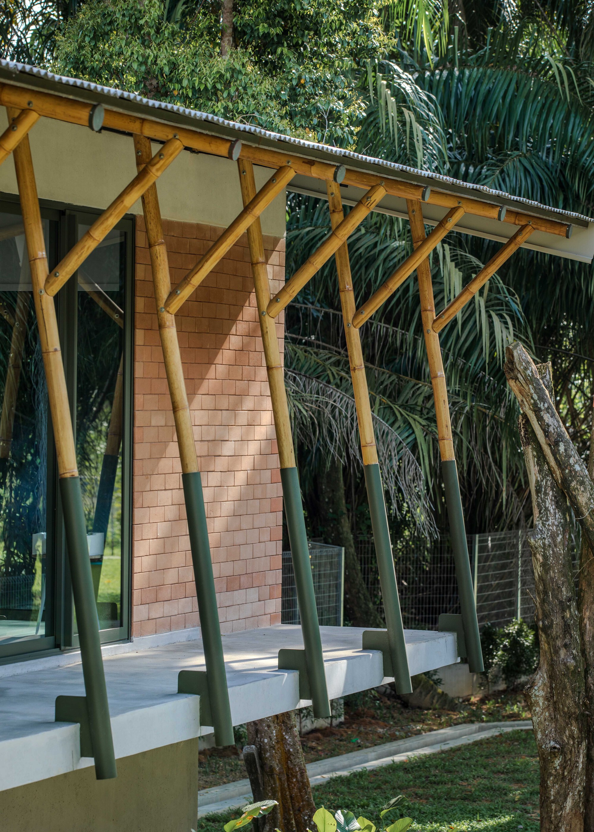 Exterior colonnade with angled bamboo columns supporting a corrugated metal roof beside lush tropical vegetation