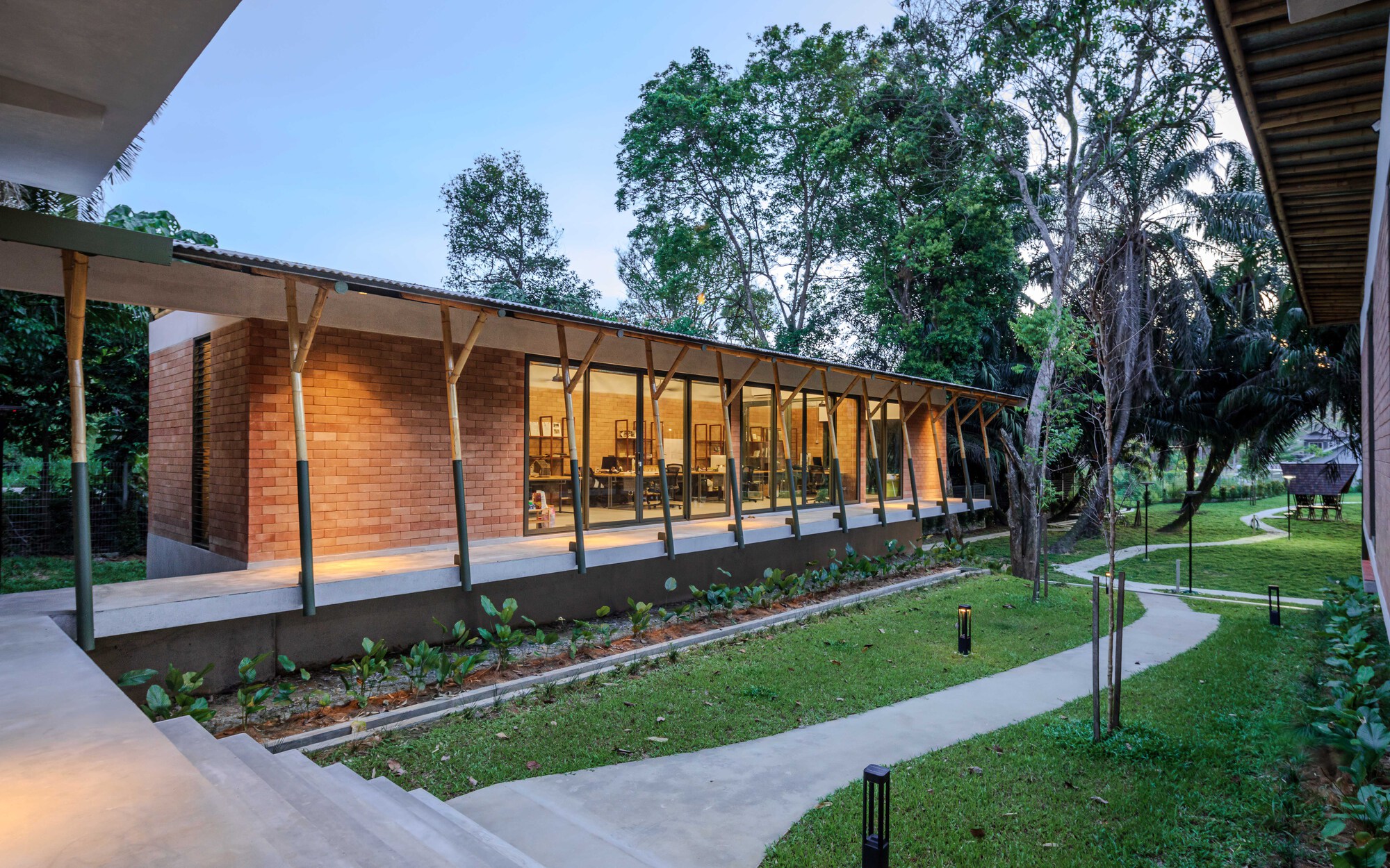 Glass-walled wing with timber-braced roof and planted bed at dusk beneath scattered trees