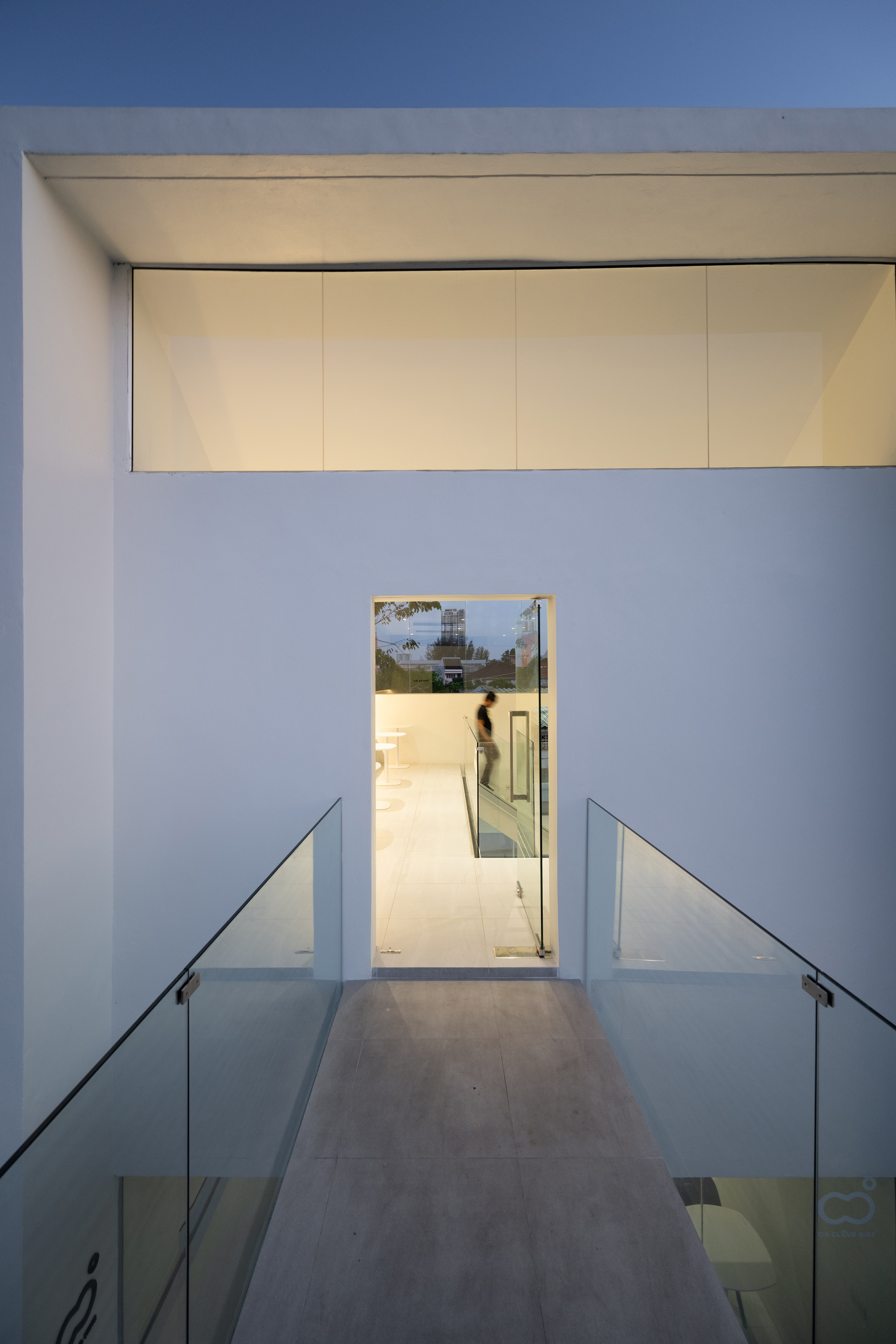 Interior passage with yellow ceiling panels and glass-railed stairs descending toward a glass door at evening