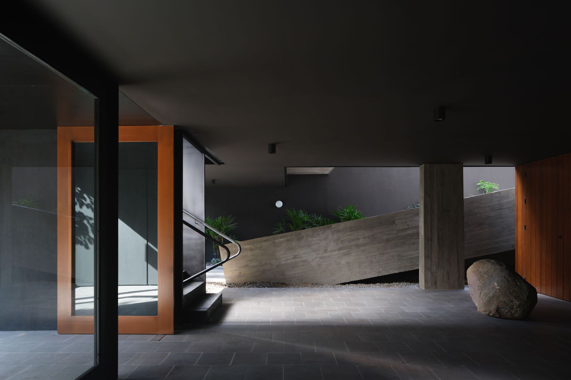 Entry foyer with angled concrete wall, timber-framed doorways casting orange light, and a large boulder