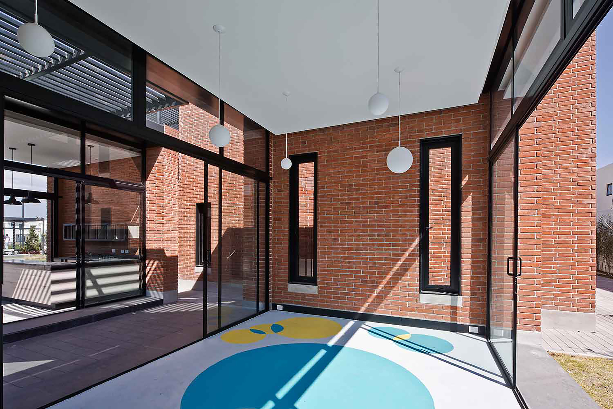 Covered brick terrace with glass walls, slim black-framed windows and a circular painted floor pattern in bright sunlight