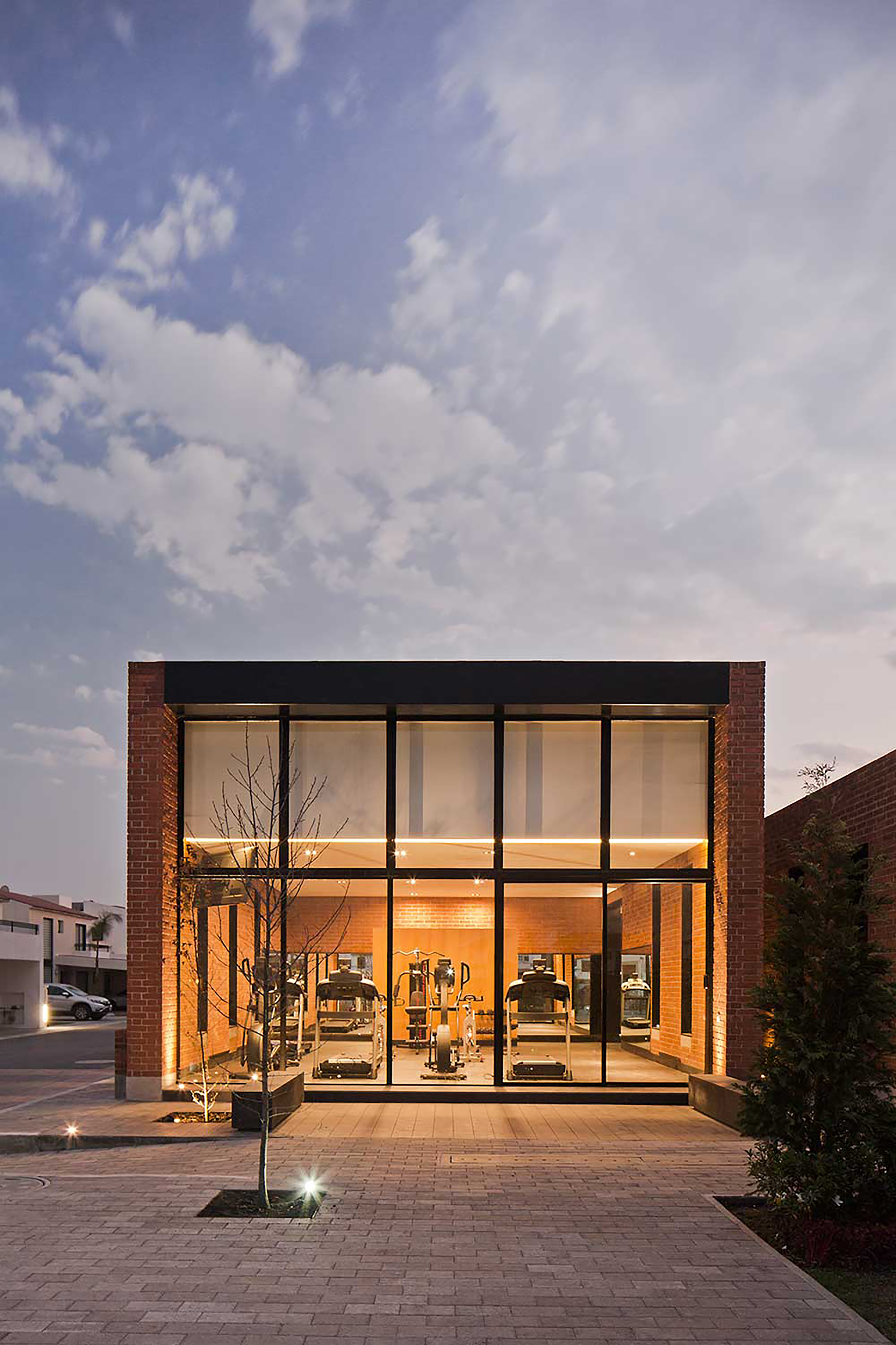 Glass-fronted gym pavilion framed by brick piers, with treadmills visible inside under a dusk sky
