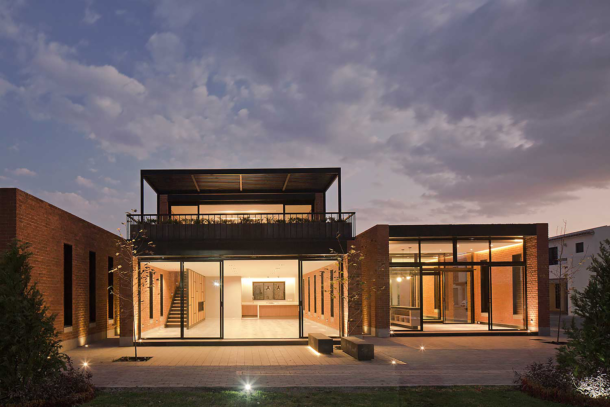 Rear view of the two-storey brick house with glazed ground floor and roof terrace, lit at dusk under heavy clouds