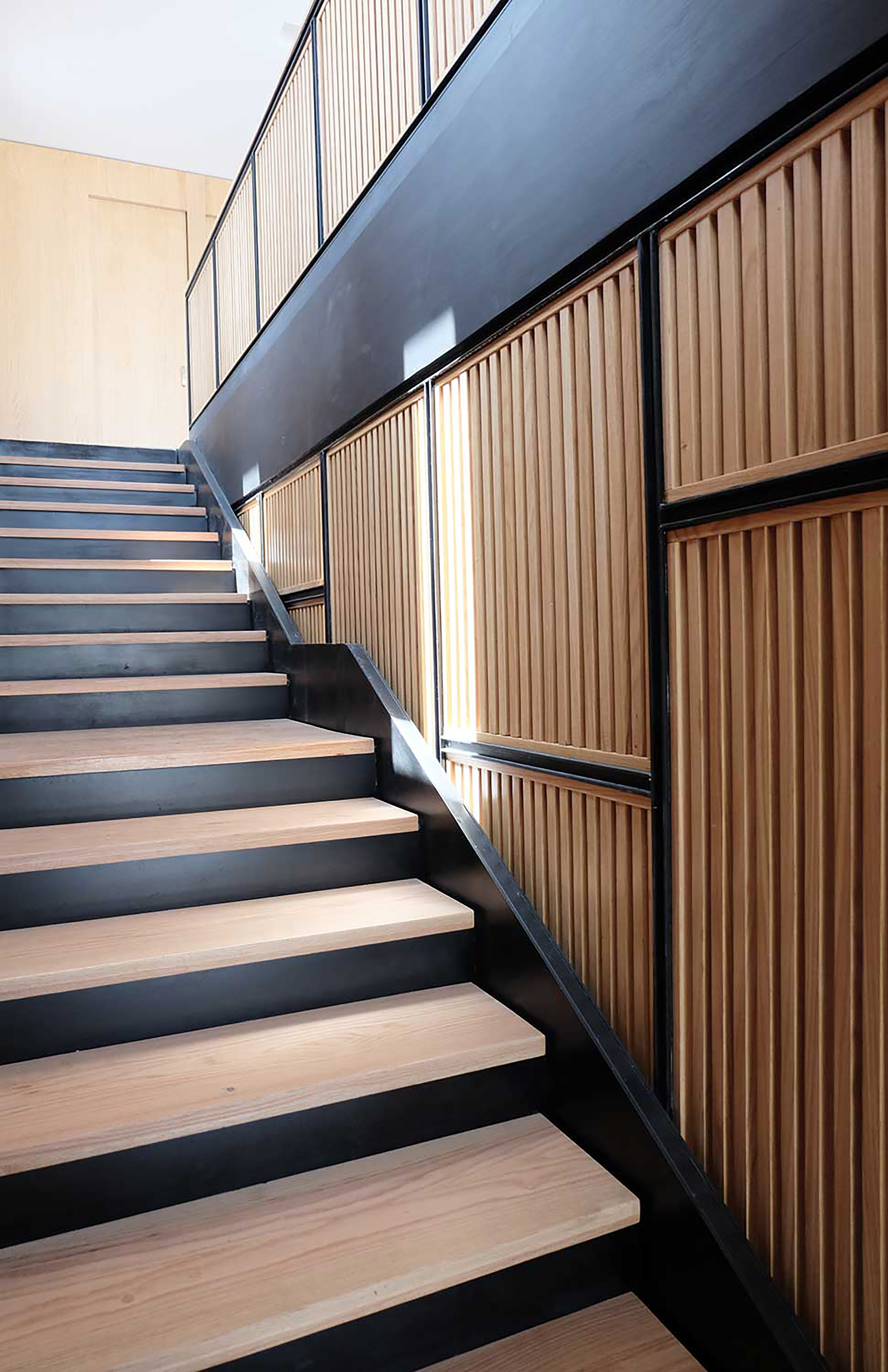 Staircase with pale timber treads, black steel stringers and vertical wood slat panels along the wall in soft daylight
