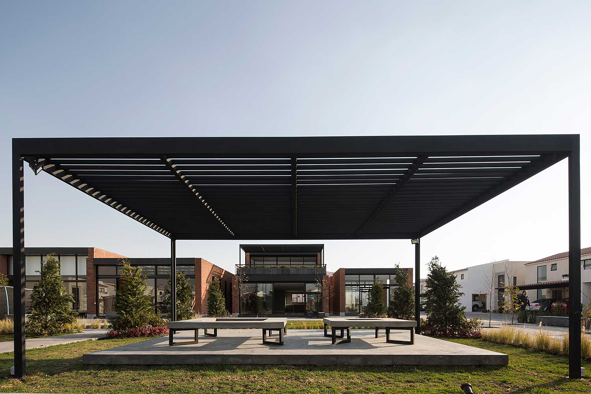 Black steel pergola with slatted roof over a concrete platform and benches, brick pavilions and young trees behind under a clear sky