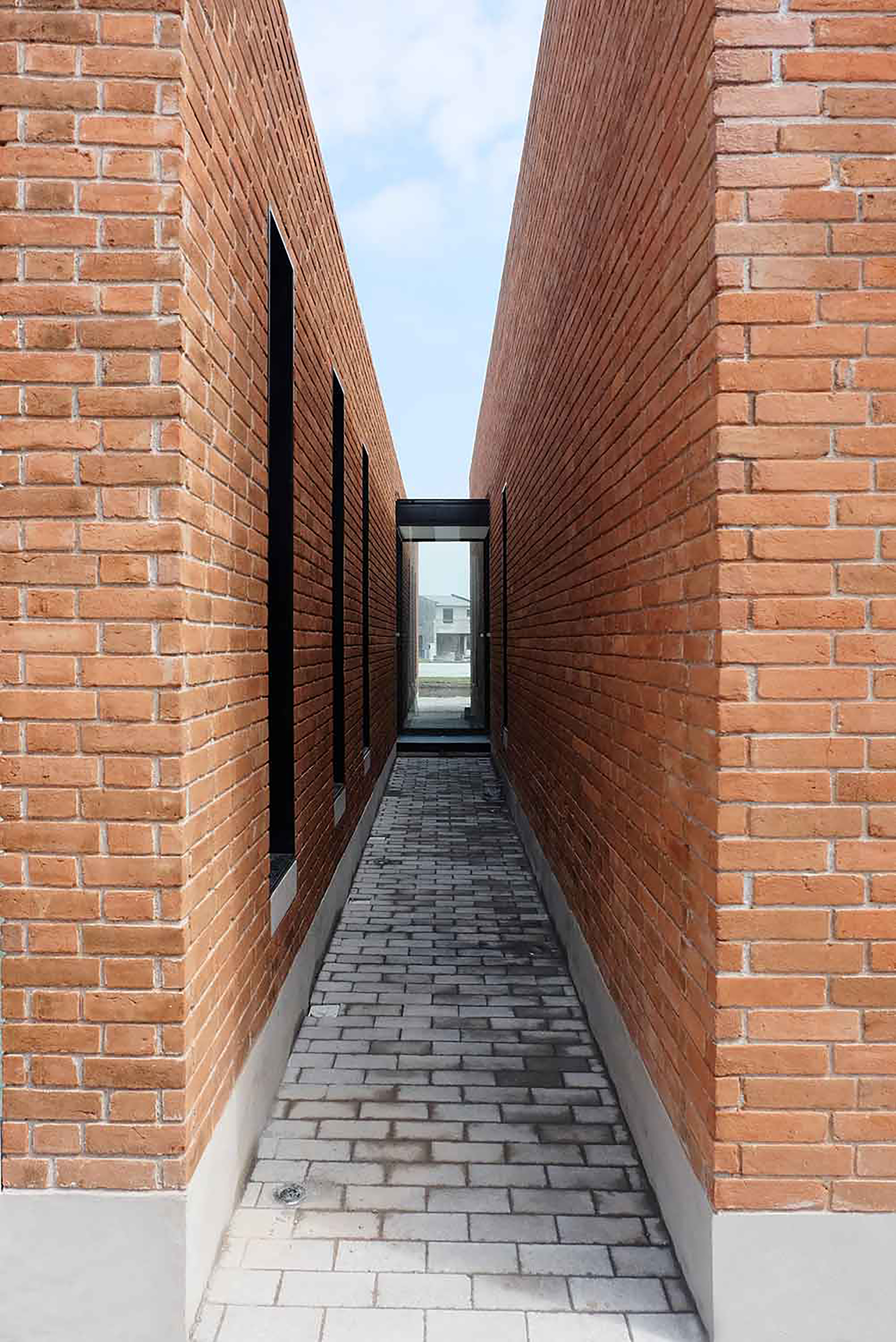 Narrow paved passage between two tall brick walls with slot windows, ending in a glazed opening under pale cloud