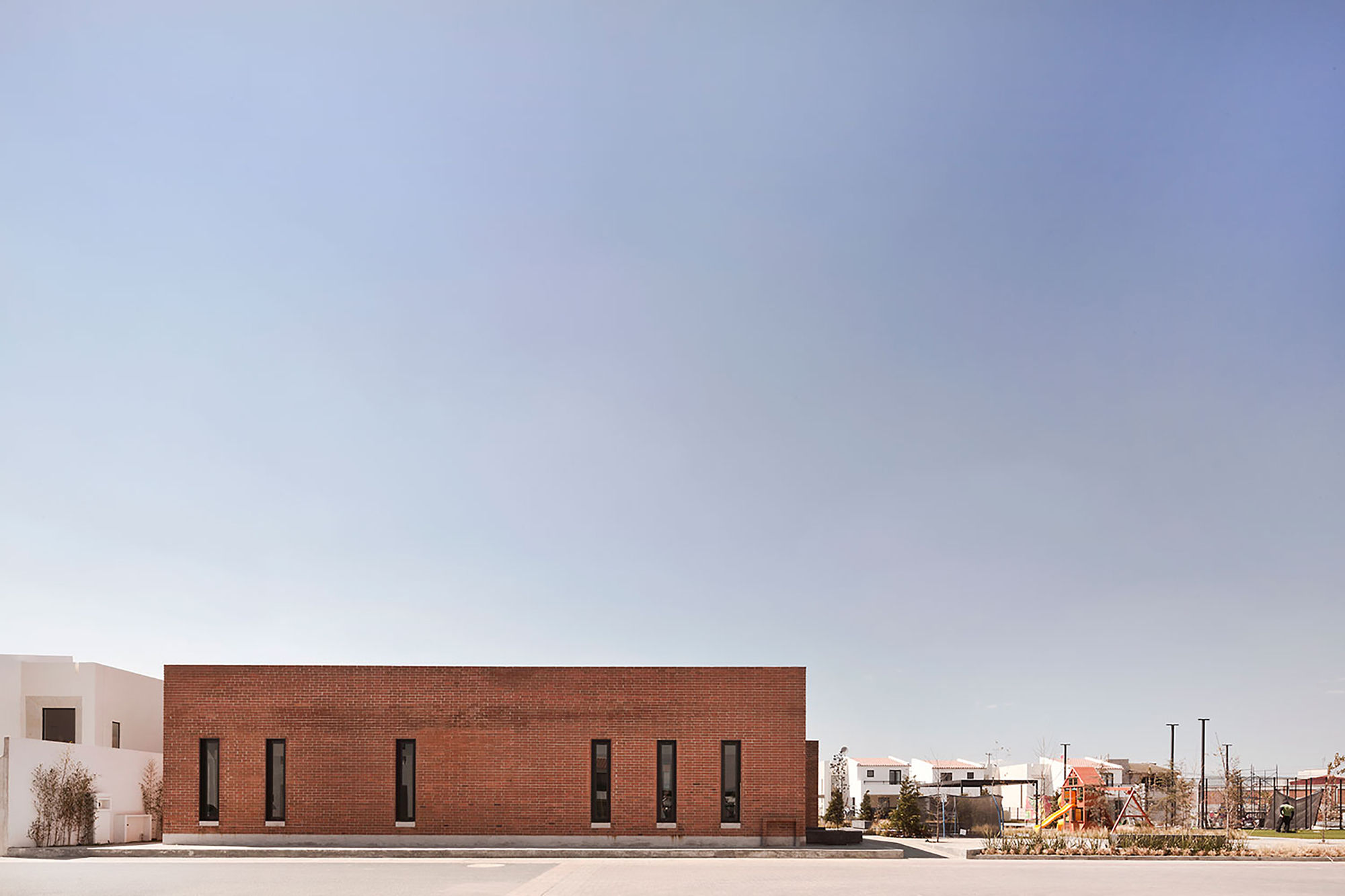 Single-storey brick volume with six tall narrow windows, a playground and white houses beyond under a hazy blue sky