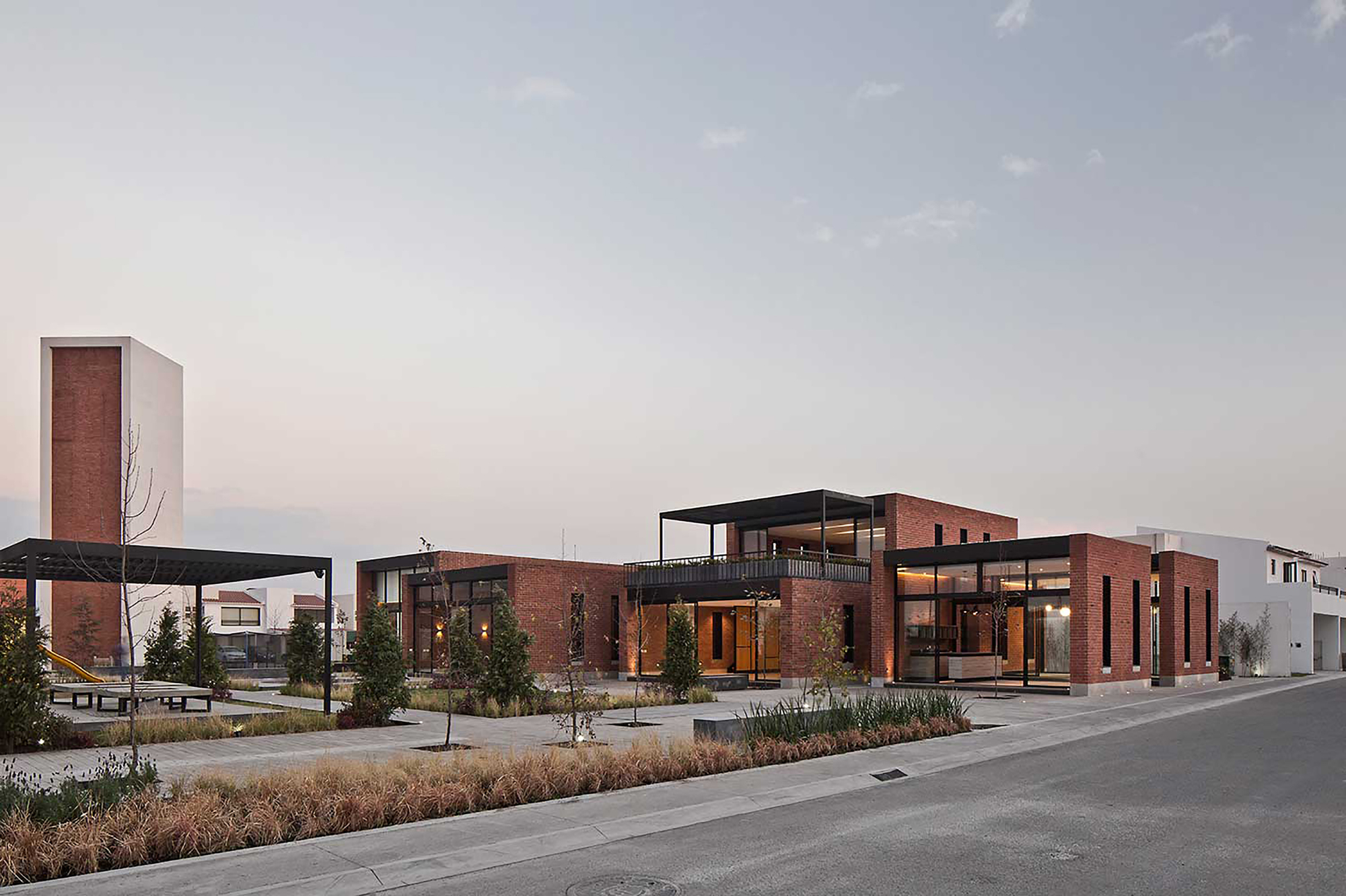 Brick pavilions with glazed corners and a roof terrace at dusk, lit interiors glowing beside a pergola and ornamental grasses