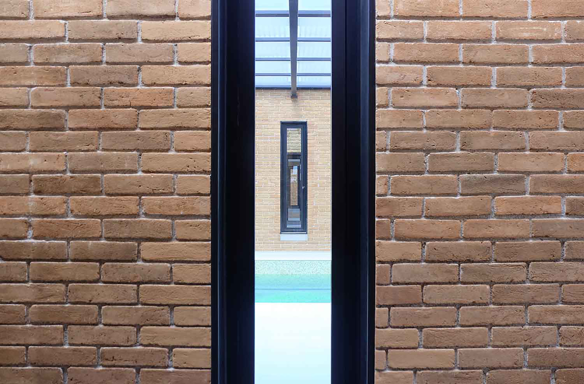 Close-up of a narrow black-framed window slot cutting through a brick wall, framing a pool and a second window beyond