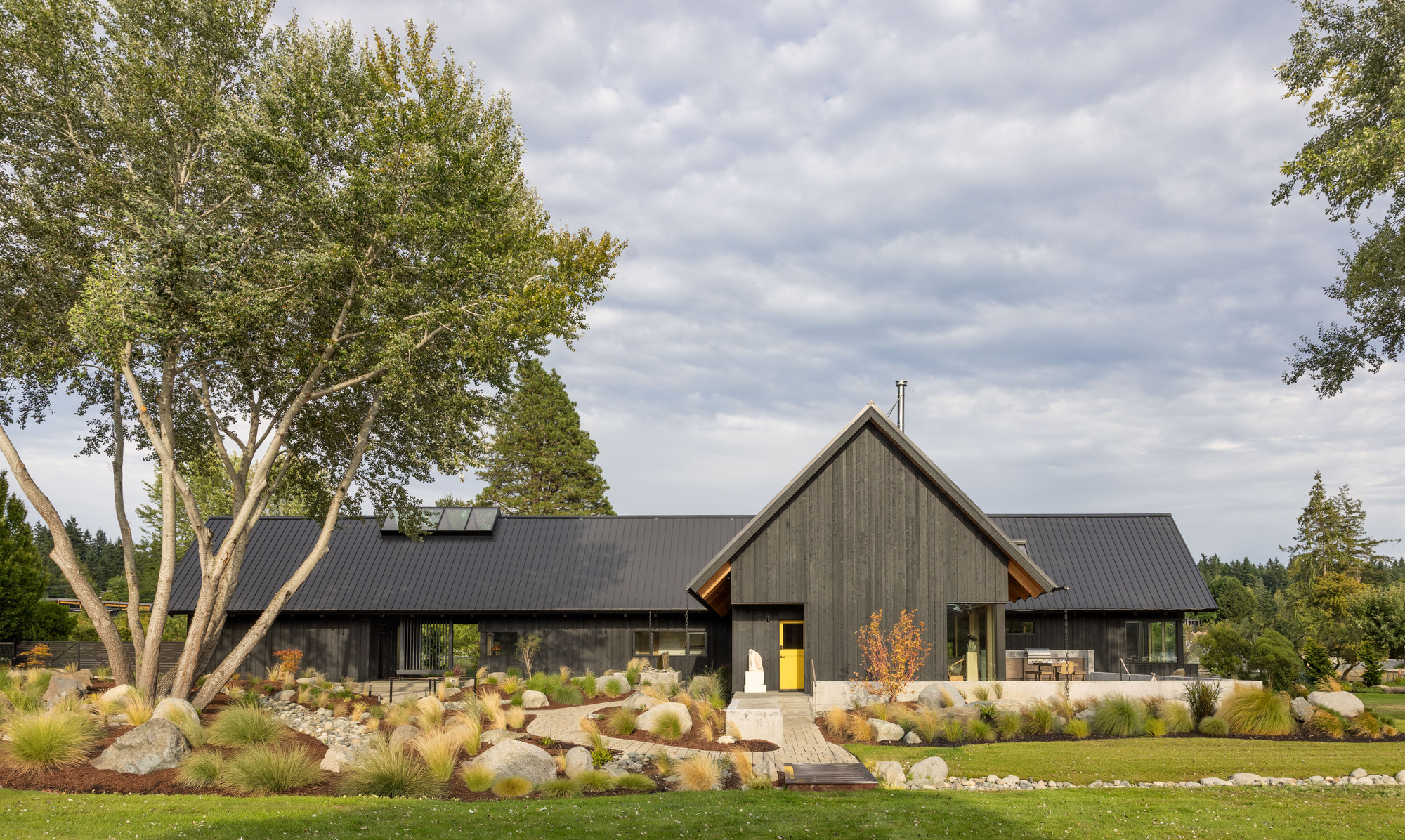 Front elevation of a charred-timber gabled entry and standing-seam metal roof behind a rock and grass garden under overcast sky