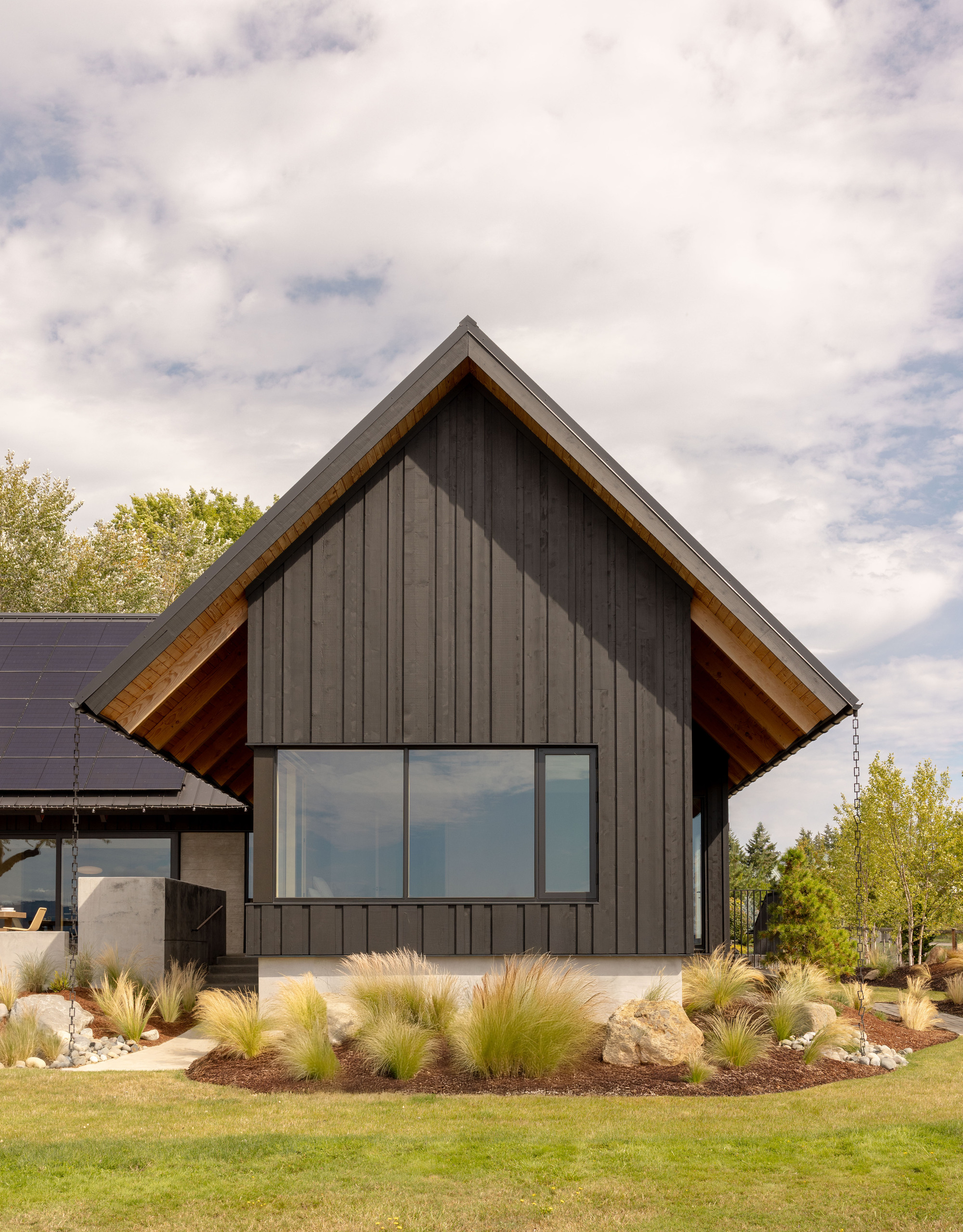 Gabled end wall of dark vertical siding with a large picture window and timber soffit, rain chains hanging at both eaves