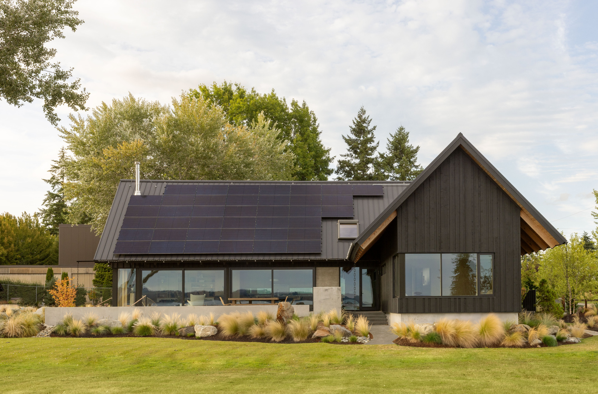 Front facade of the dark timber-clad house with a gabled entry and rooftop solar panels, set behind ornamental grasses under soft cloud