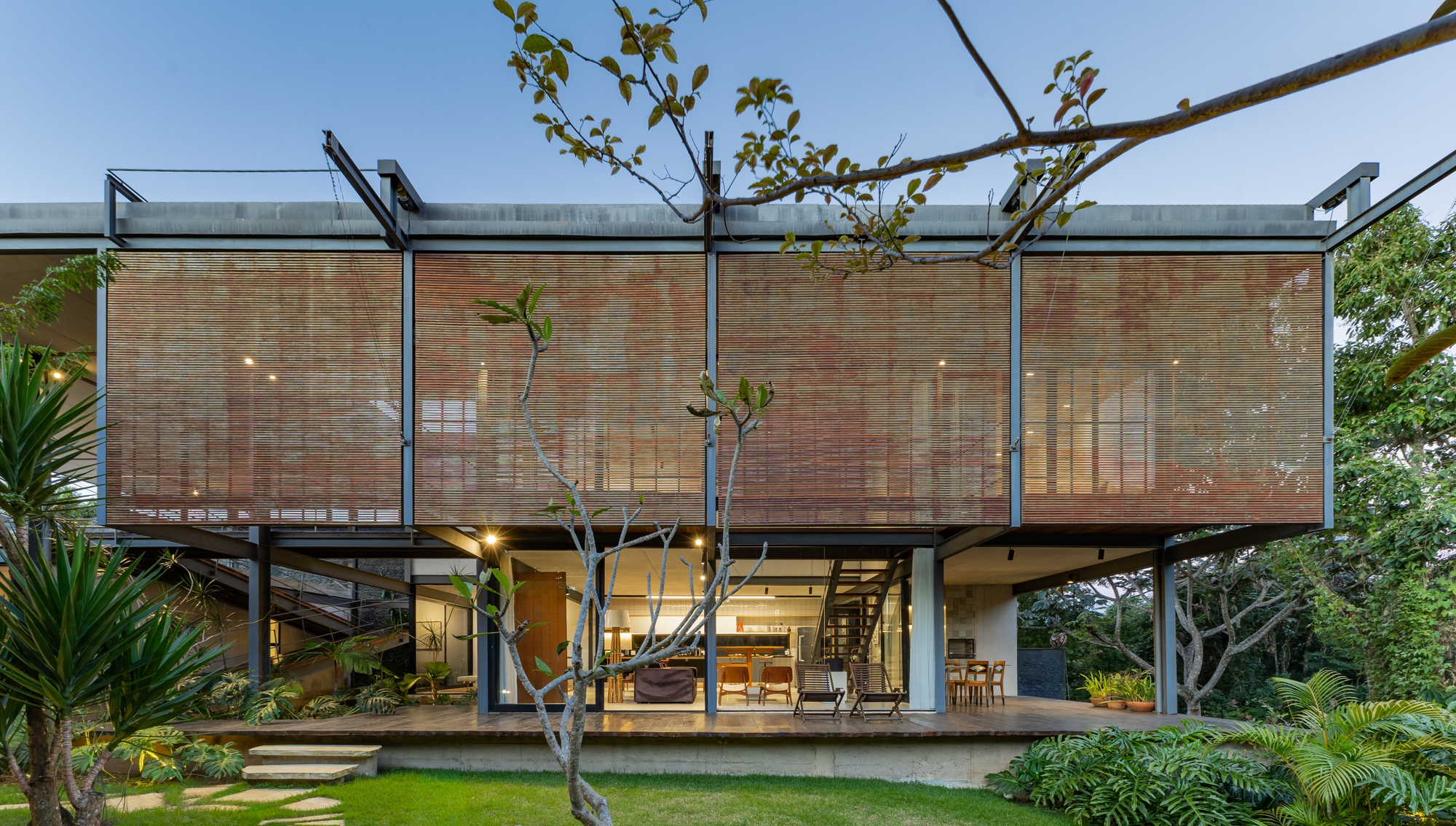 Garden facade with raised bamboo-slat screens over a glazed living level, seen at dusk through frangipani branches and lawn
