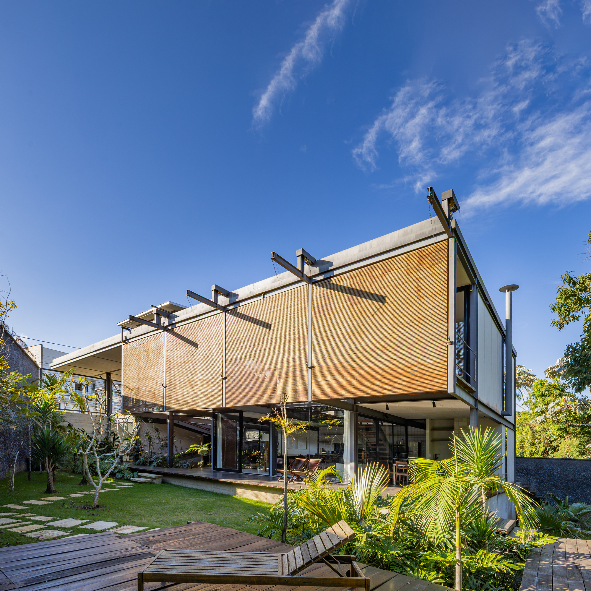 Corner view of the steel-framed house with timber slat screens, a deck lounger and palms under a blue sky with wispy clouds