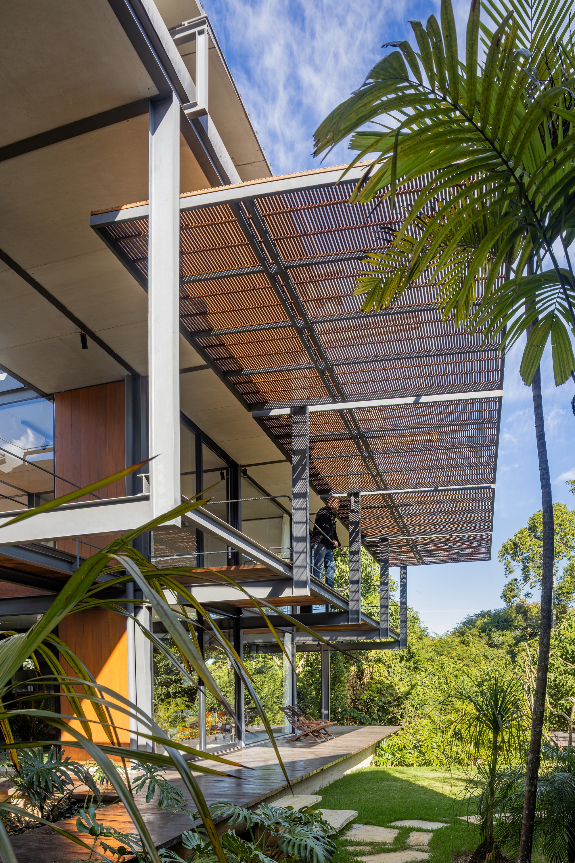 Slatted timber canopy cantilevering from steel columns above a wooden deck, with a person standing on the upper walkway and a palm in front