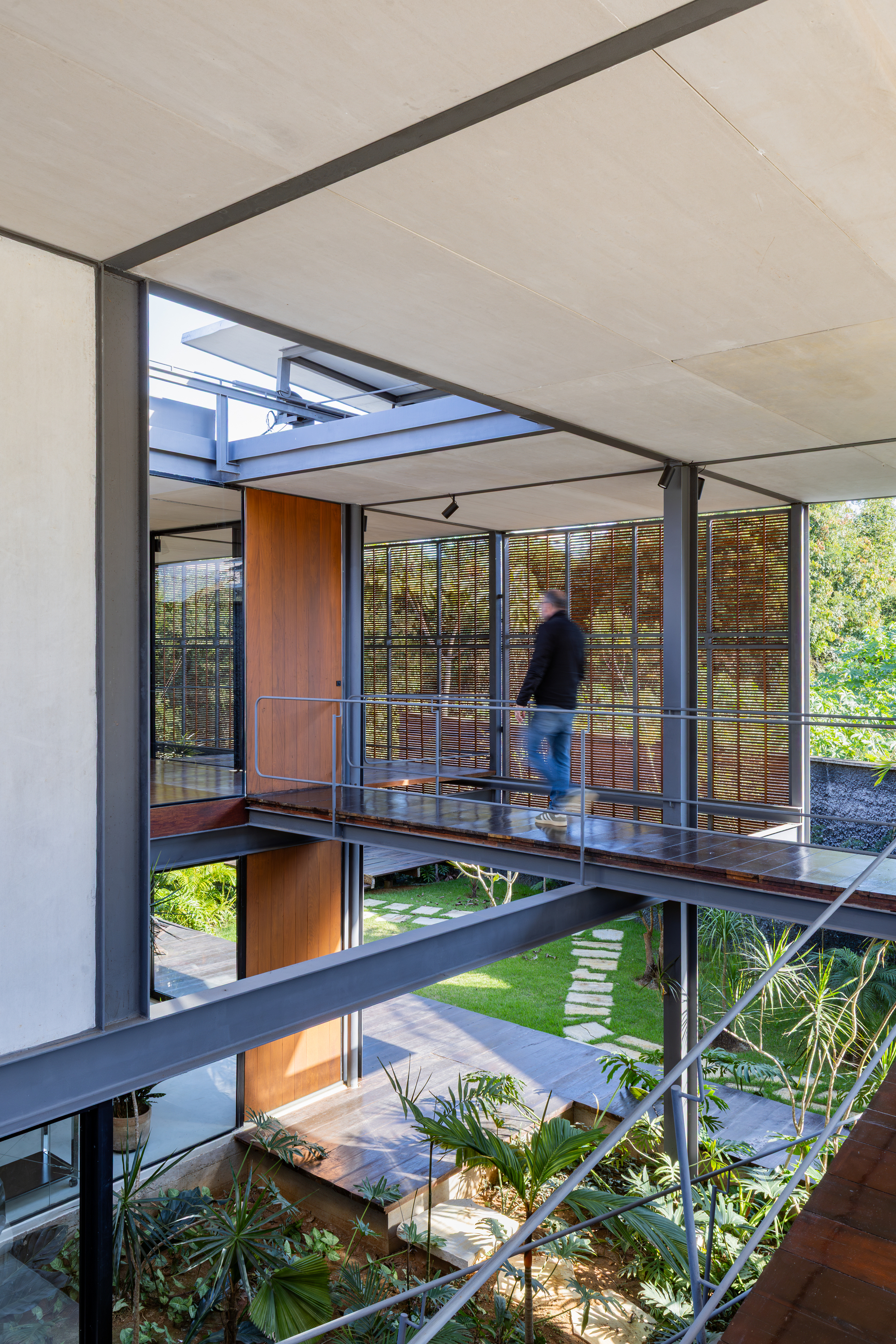 Person crossing a timber-decked steel bridge between glazed volumes, with a stepping-stone path and planted courtyard below in afternoon light