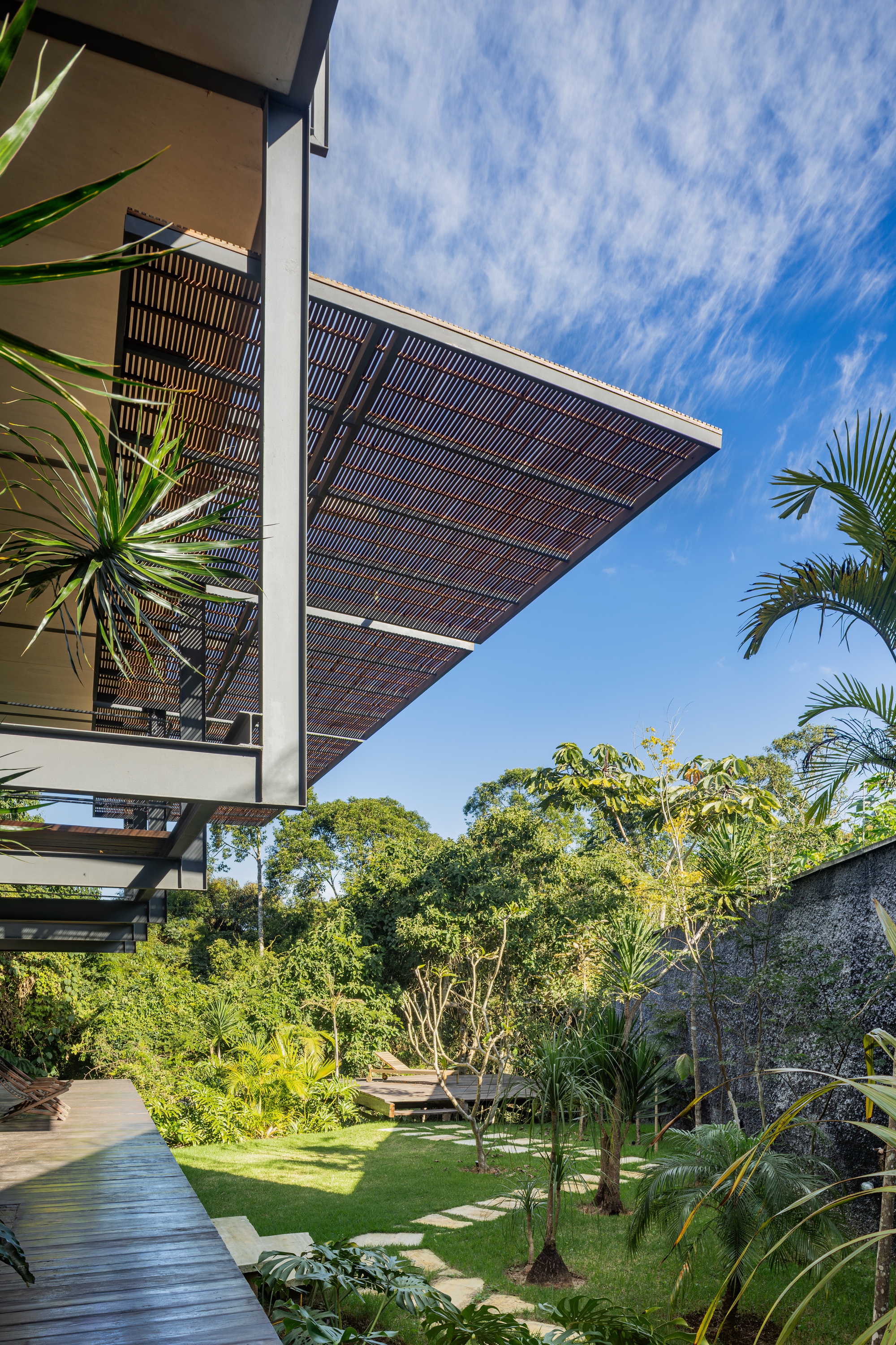 Underside of the cantilevered slatted canopy on steel framing, above a lawn with stepping stones, a stone wall and wispy clouds