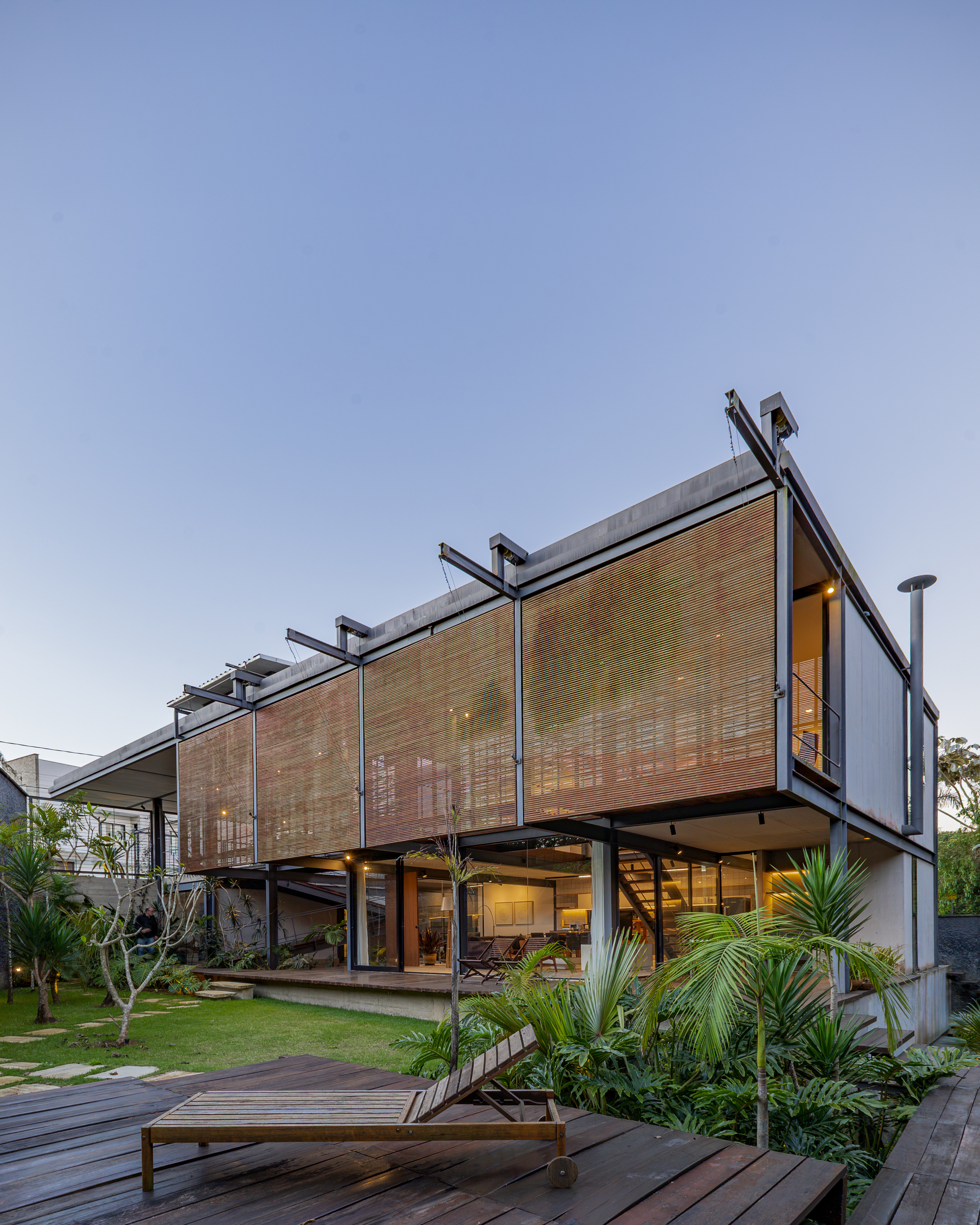 Two-storey steel-framed house with raised timber slat screens glowing at dusk, a wooden lounger on the deck in the foreground