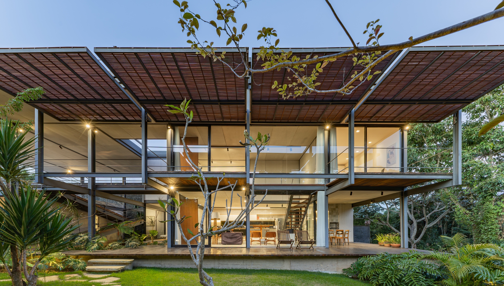 Full-width view of the steel frame with overhanging woven canopy and glazed living level, lit from within at blue hour