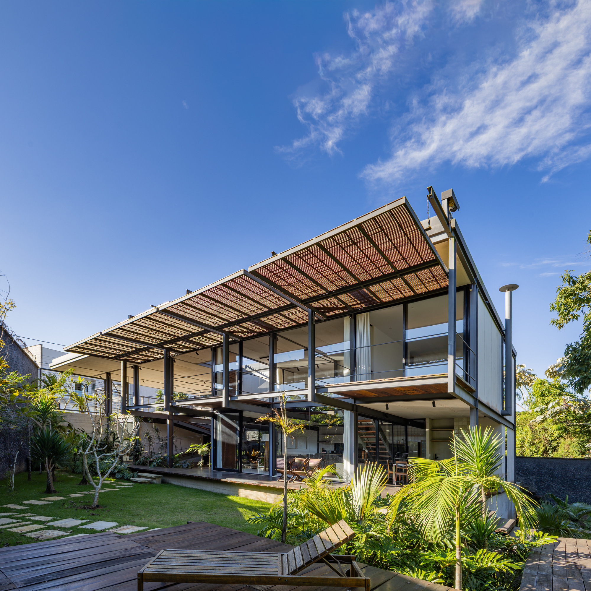 Steel and glass house with a tilted woven canopy over the upper floor, seen across lawn and stepping stones under blue sky