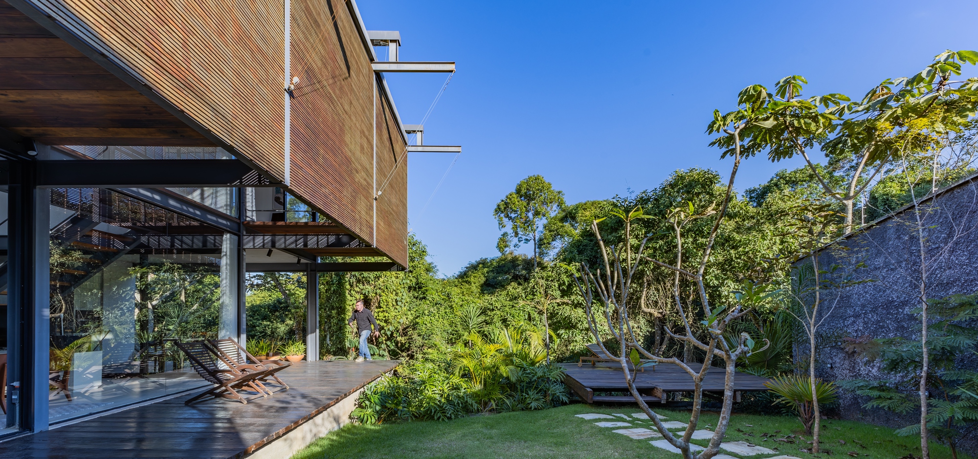 Side terrace with timber deck and loungers beneath the slatted upper volume, a person walking near the garden wall in daylight