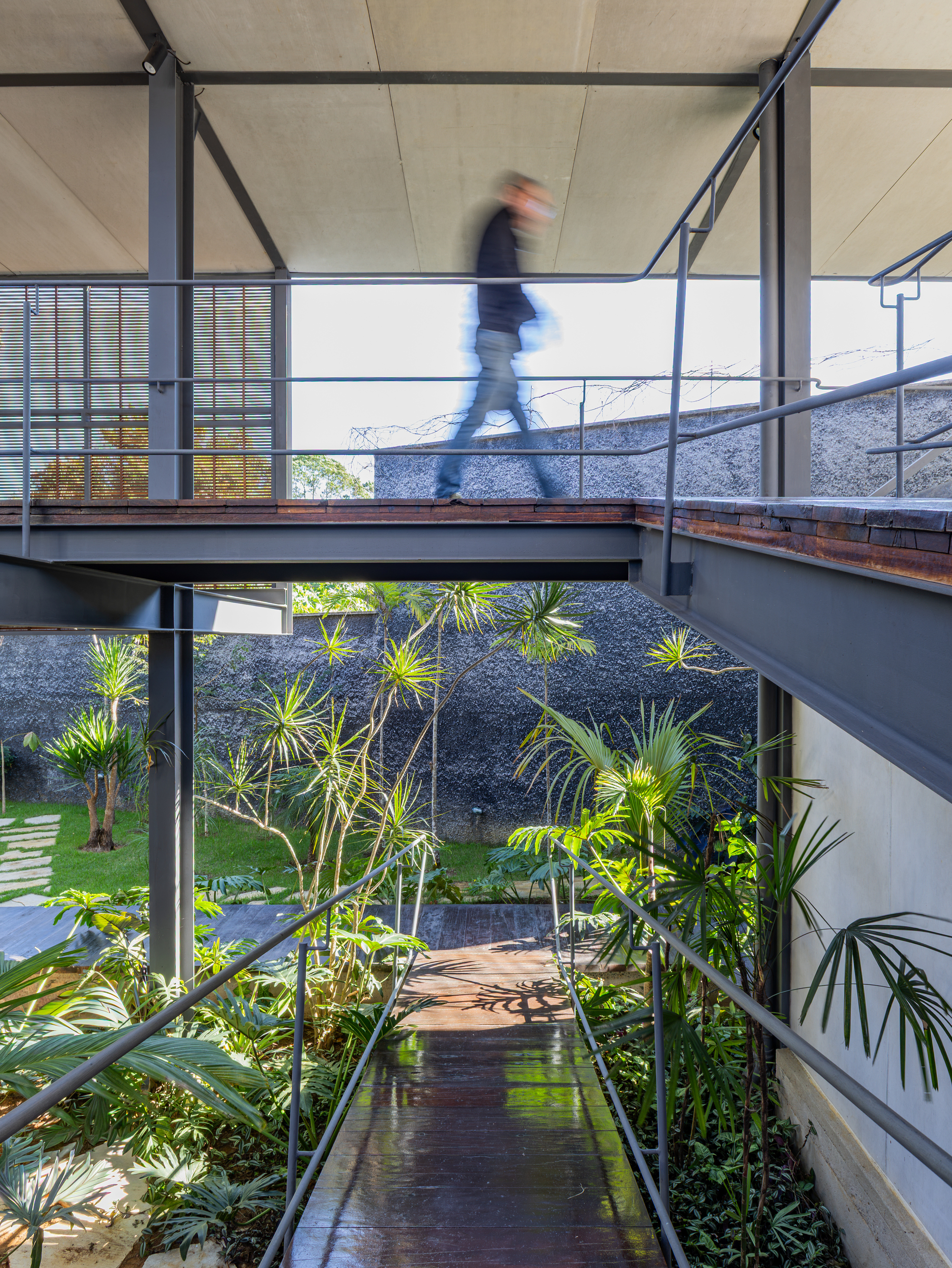 Person in motion crossing a steel-framed timber footbridge above a planted courtyard with palms, beside a dark stone wall in daylight