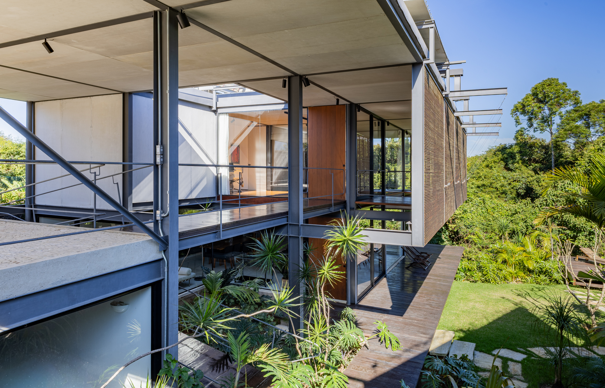 Steel-framed house volumes with timber decks and a slatted screen, set over a palm garden under a clear blue sky