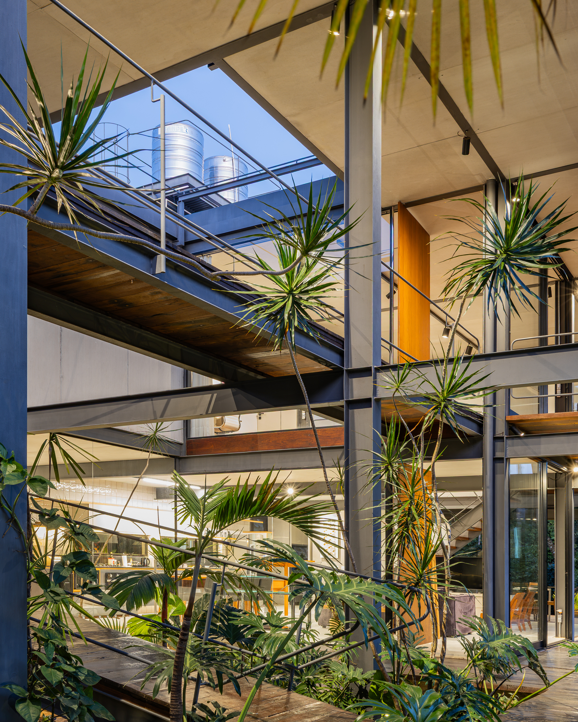 Steel columns and beams crossing a palm-filled courtyard at dusk, with warm interior light and rooftop water tanks above