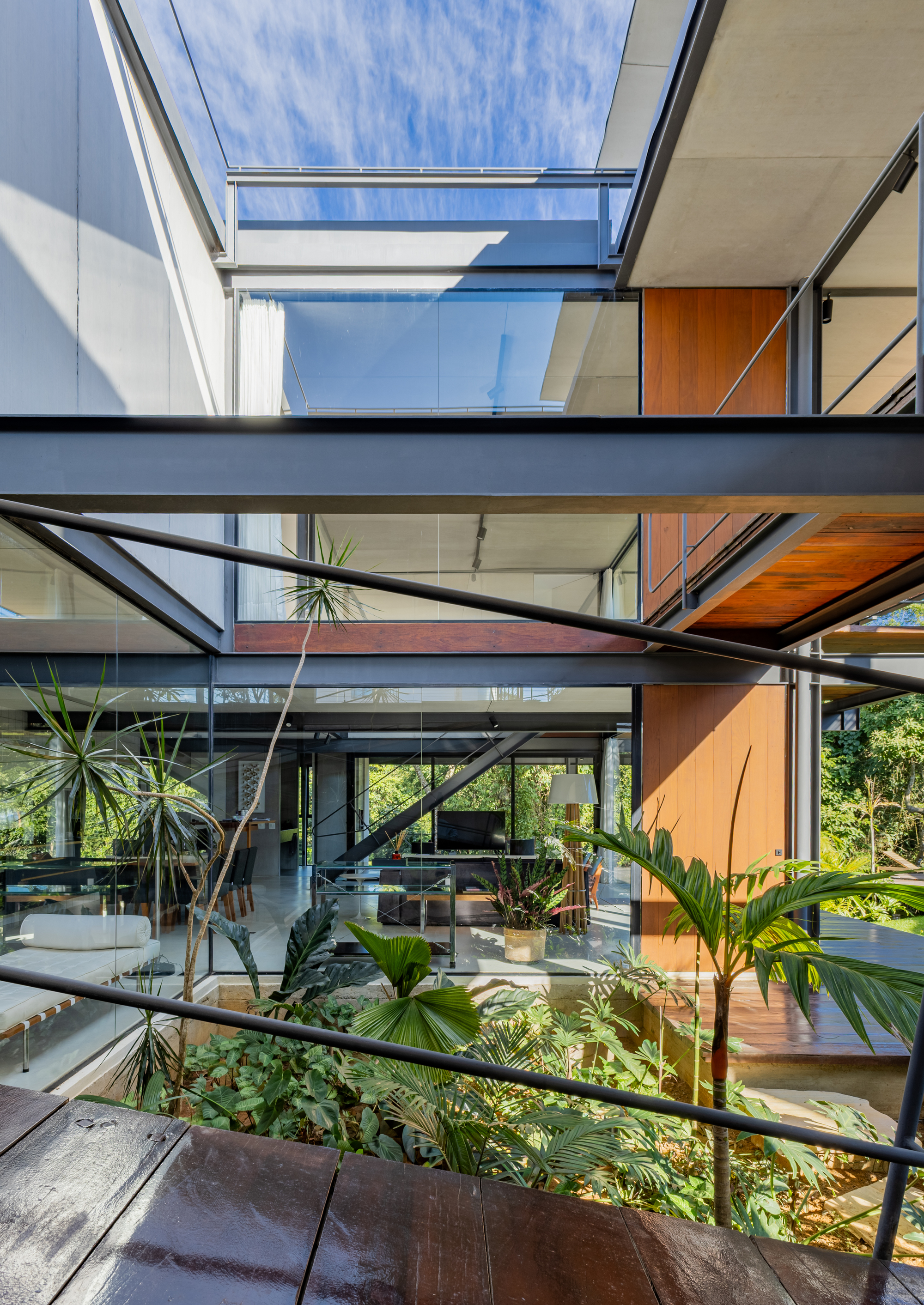 Looking across a timber walkway to stacked steel beams, glazed living space and wood-clad panels, with palms below and cloud overhead