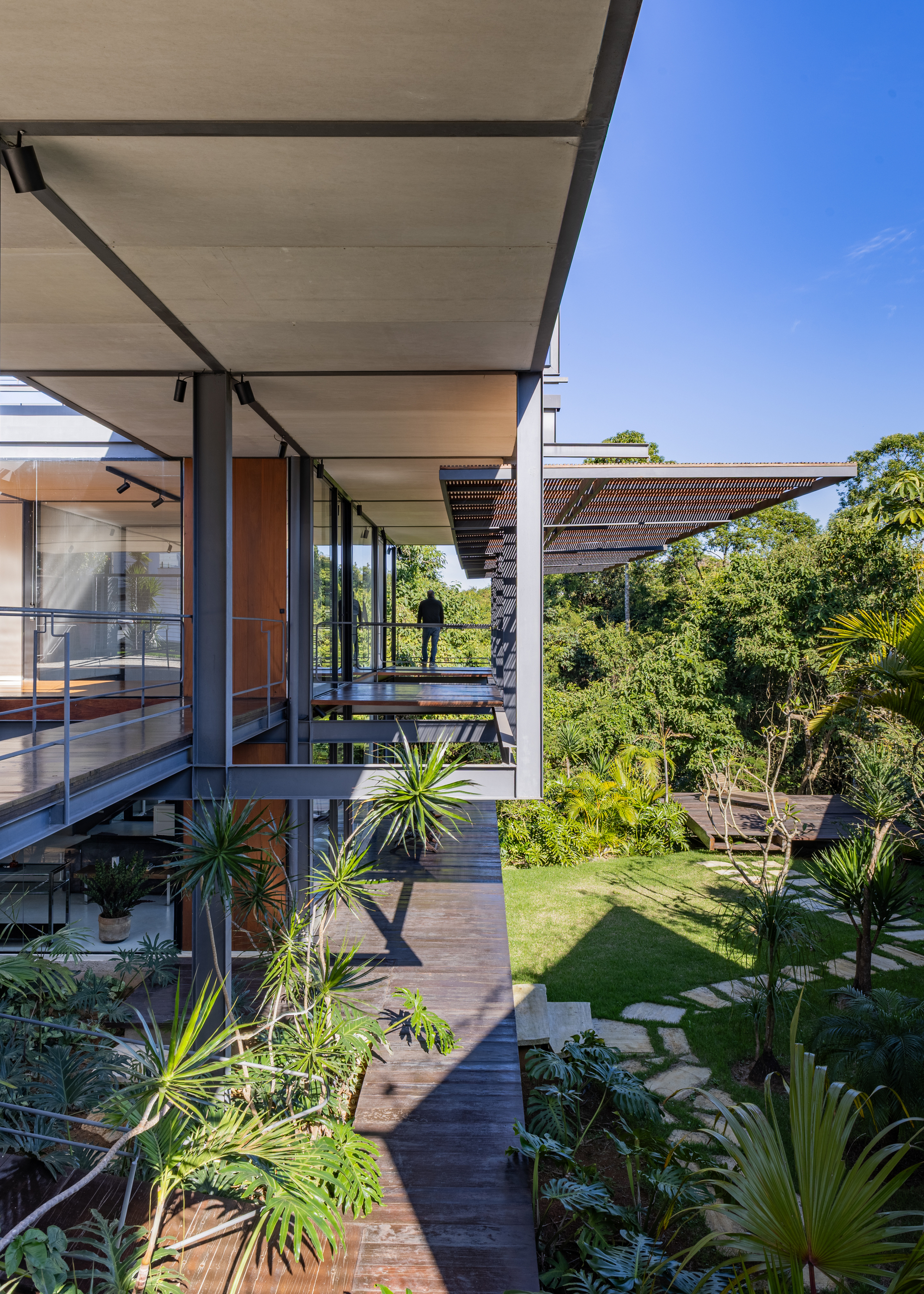 Timber deck path running beneath a wide flat roof on steel columns, with a person standing at the far railing in sunlight