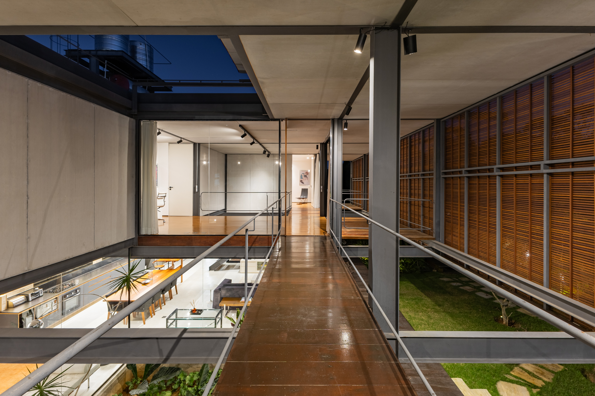 Steel-railed timber bridge crossing between glazed rooms and louvered wooden screens, seen at dusk over a lawn and living area below