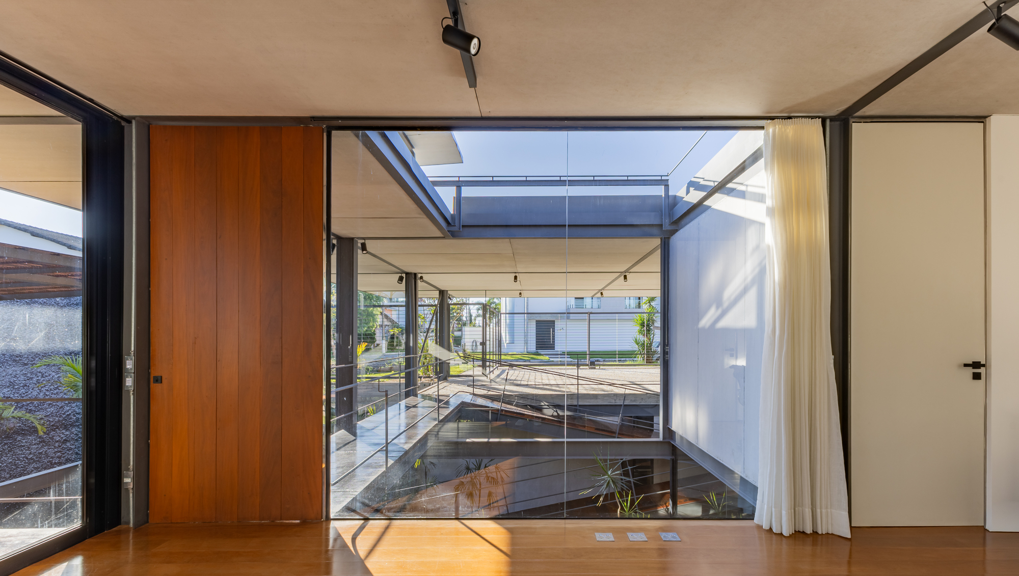 Interior view through a floor-to-ceiling glass wall toward a courtyard with a walkway, framed by a timber panel and white curtain