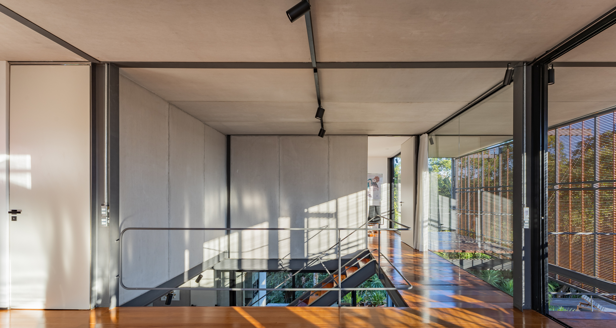 Upper-floor landing with a stair opening, concrete panel walls and track lights, with afternoon sun falling across the timber floor
