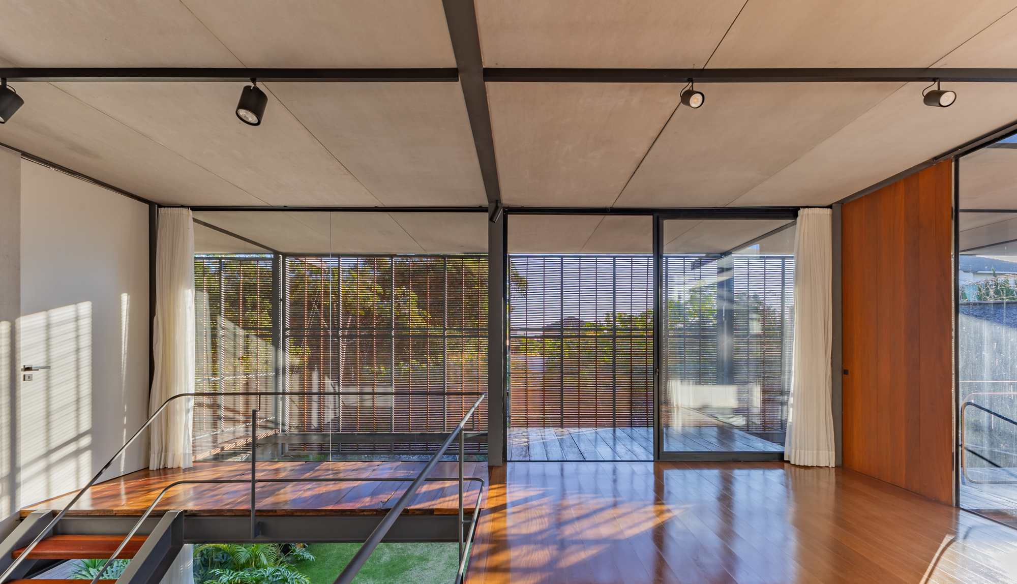 Timber-floored room with steel ceiling beams and white curtains opening toward louvered screens, with low sun casting patterns on the floor