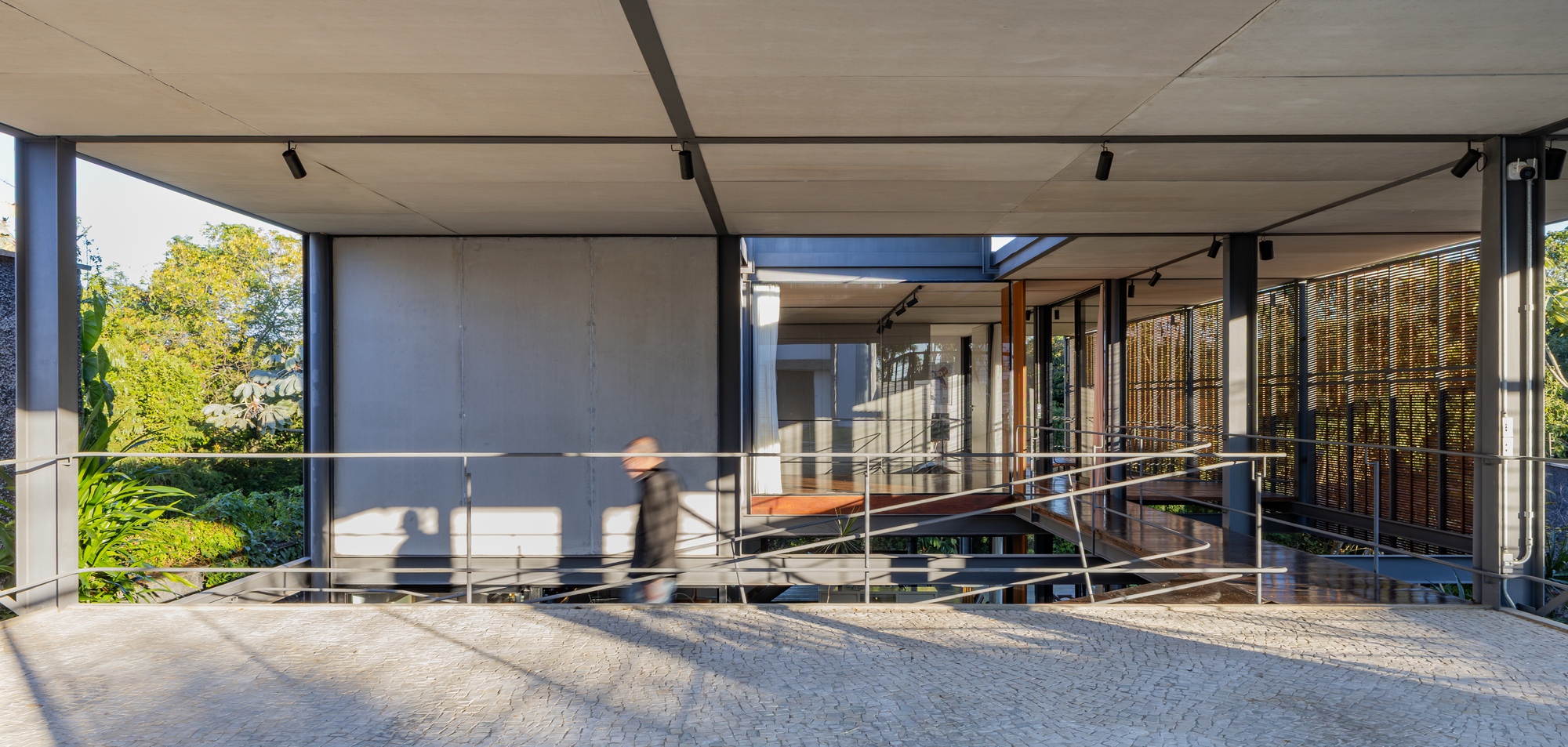 Ground-level view of the steel-framed pavilion with concrete panels and a blurred passerby walking along the stone-paved edge in sunlight