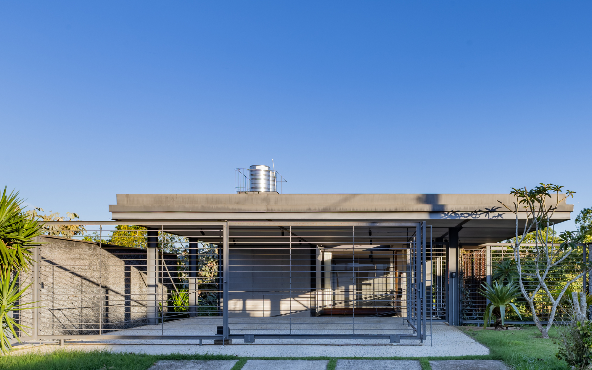 Street-side view of the flat-roofed steel pavilion with horizontal bar screens and a water tank, under a clear blue sky