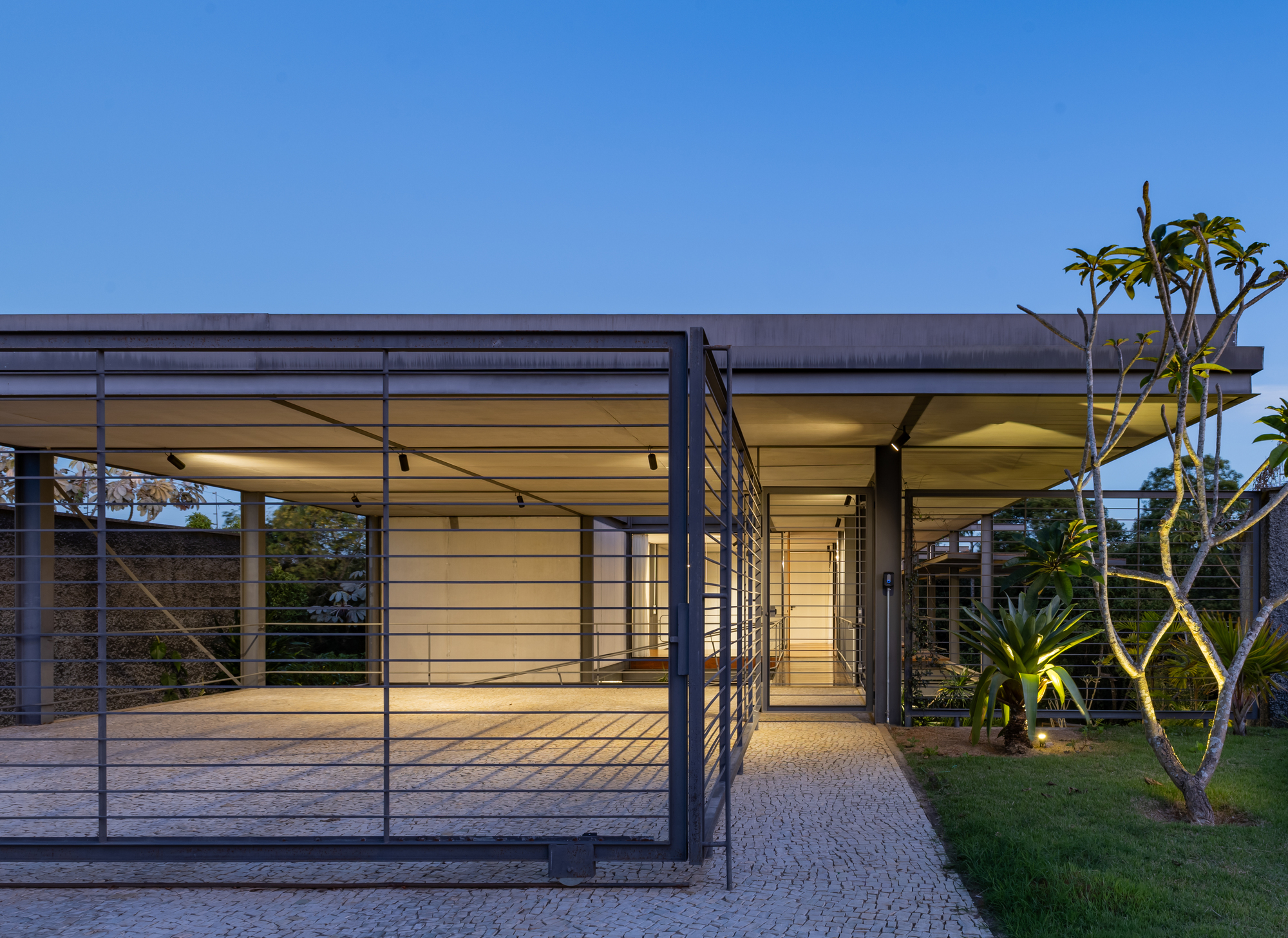 Cobblestone path leading to the open carport and entry beneath a thin flat roof at dusk, with a frangipani tree beside it