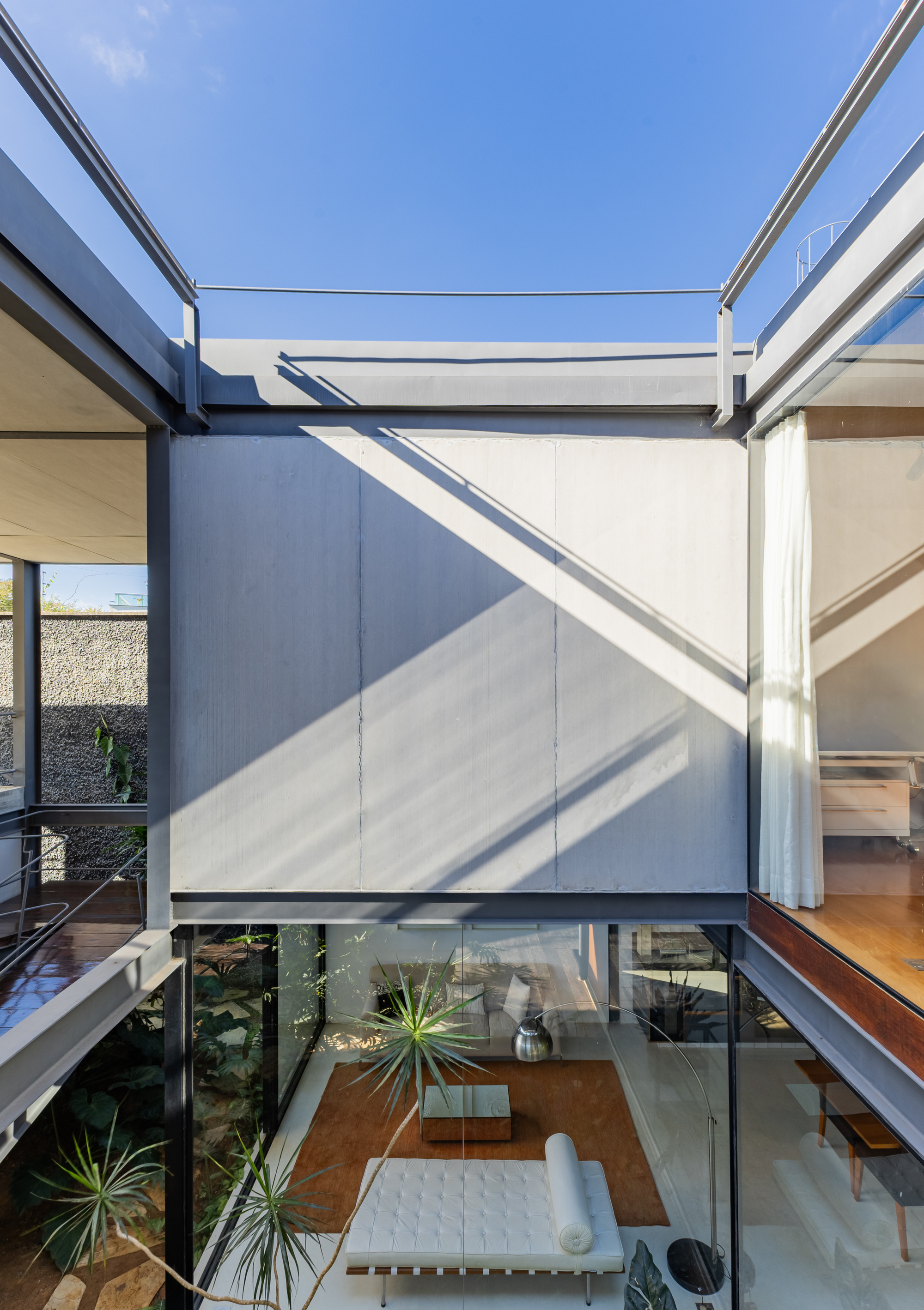 Looking up through the open courtyard to a concrete panel wall and glazed living room with a white daybed, in strong sunlight