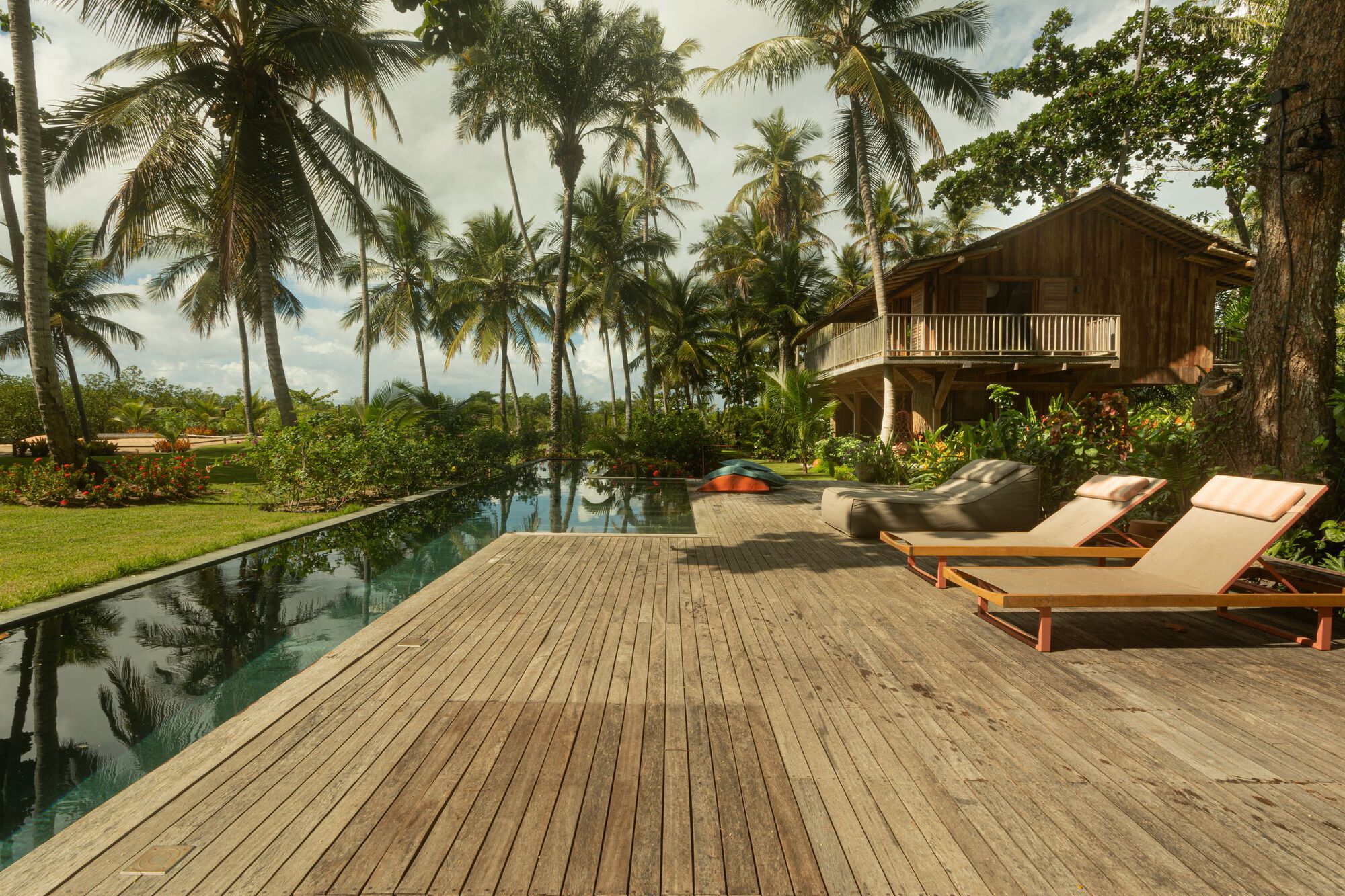 Long weathered timber pool deck with two lounge chairs beside a narrow reflecting pool and palms under scattered clouds