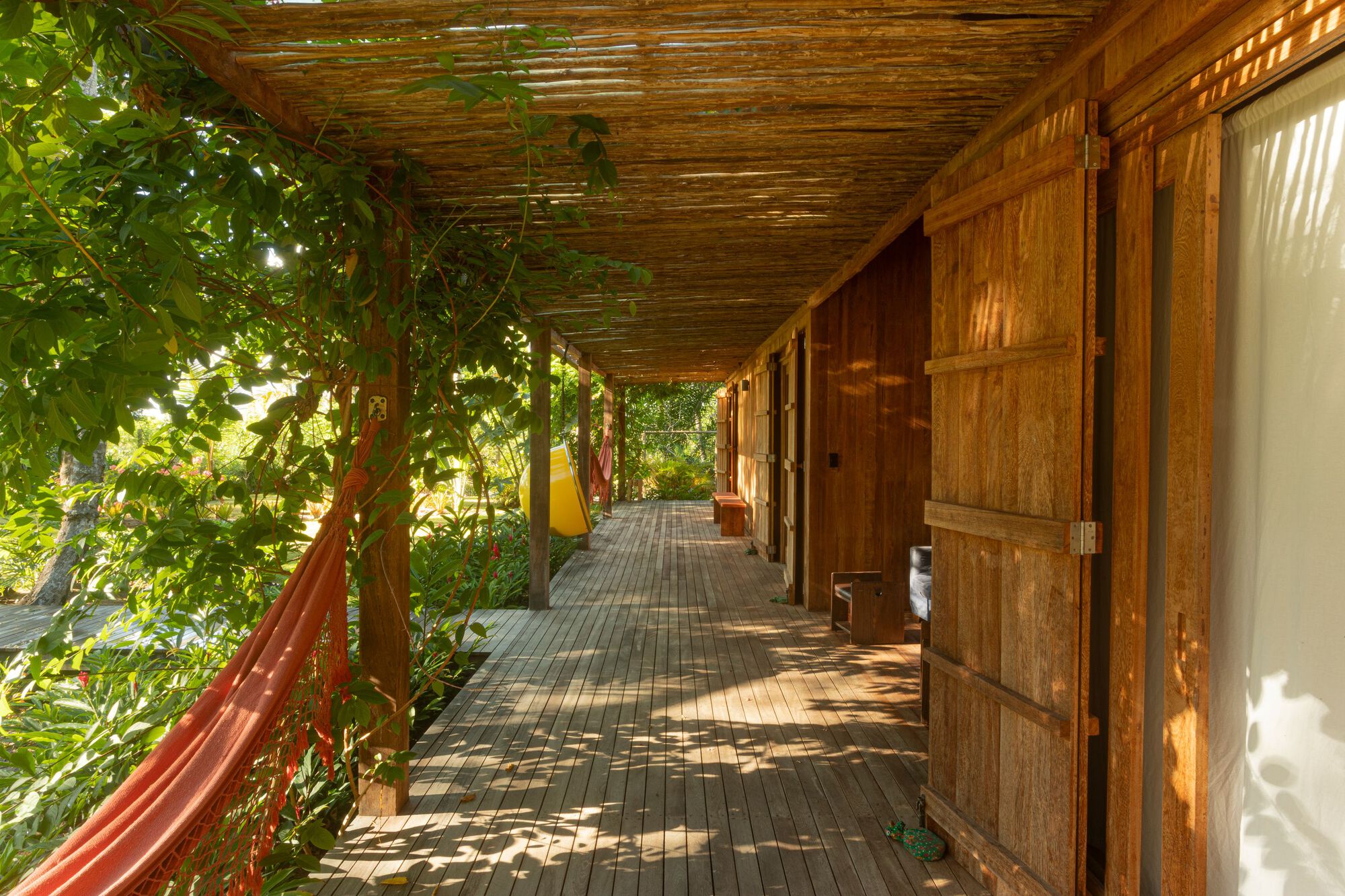Covered timber veranda with woven cane ceiling, plank shutters and a red hammock, dappled by leaf shadows