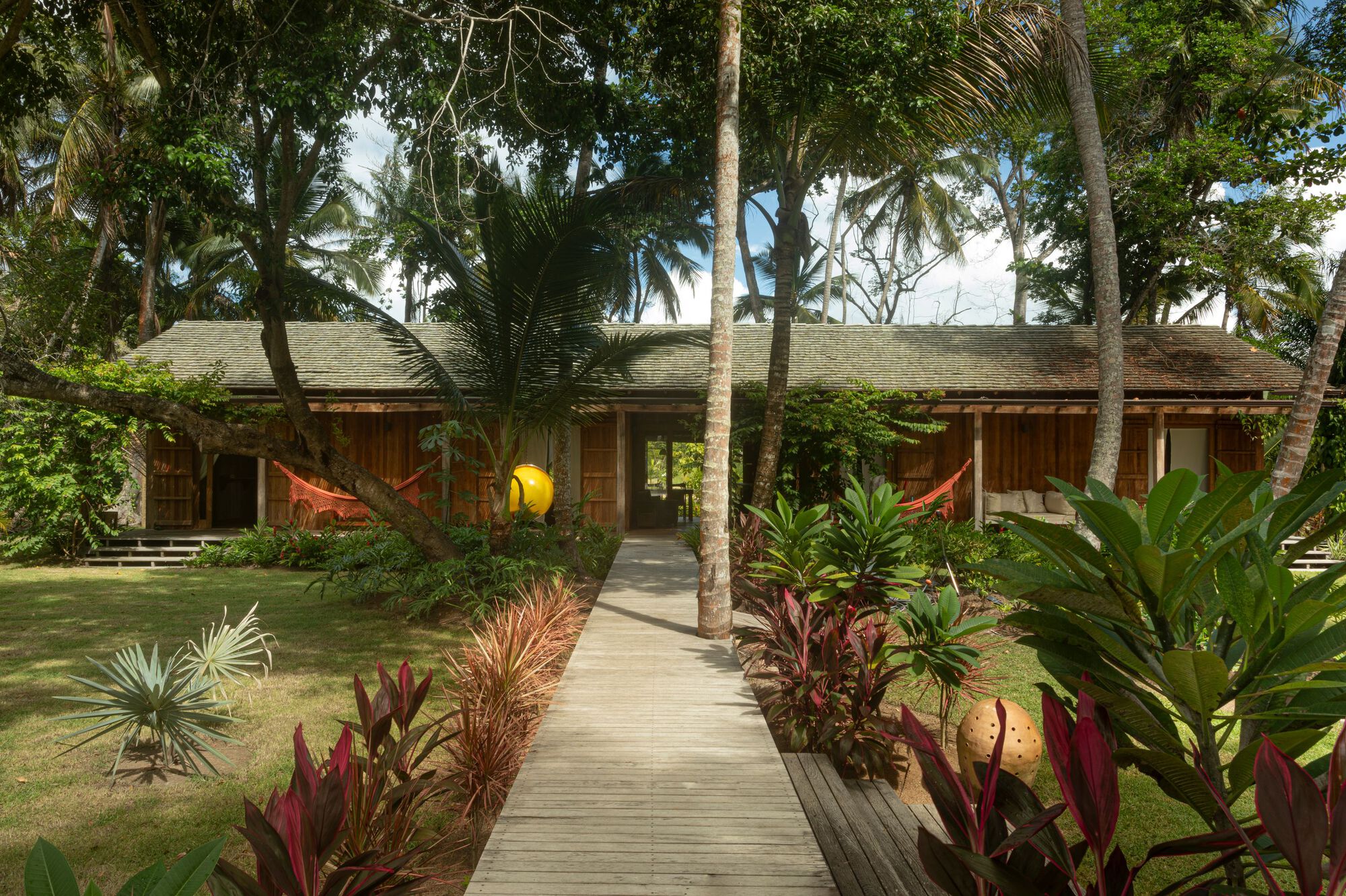 Low timber pavilion with a shingle roof reached by a plank boardwalk through palms and red-leaved plants
