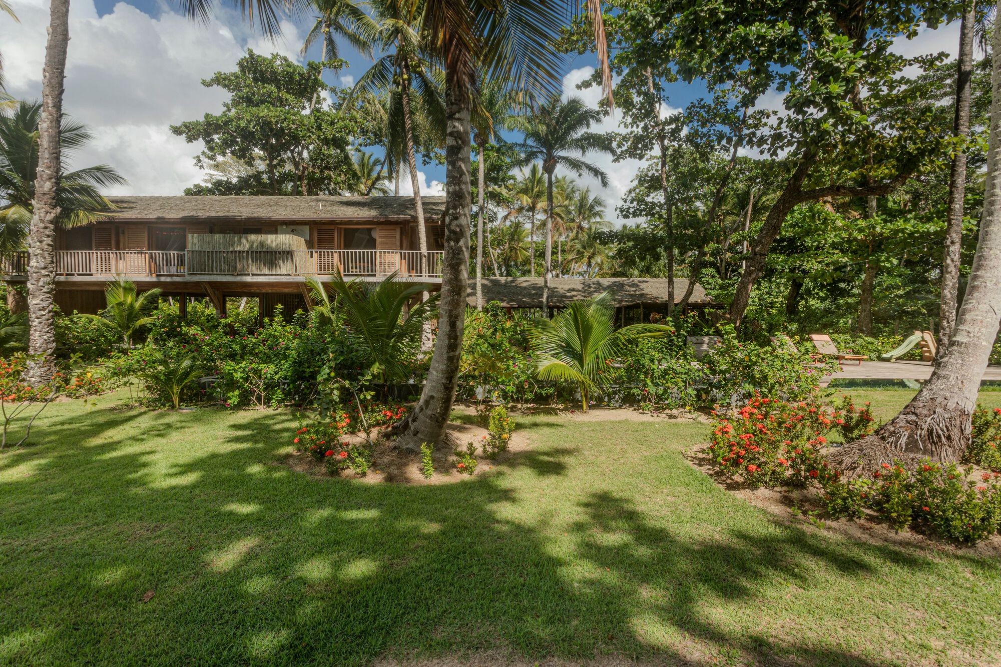 Two-storey timber house with a railed upper balcony seen across a lawn striped with palm shadows and red flowering shrubs