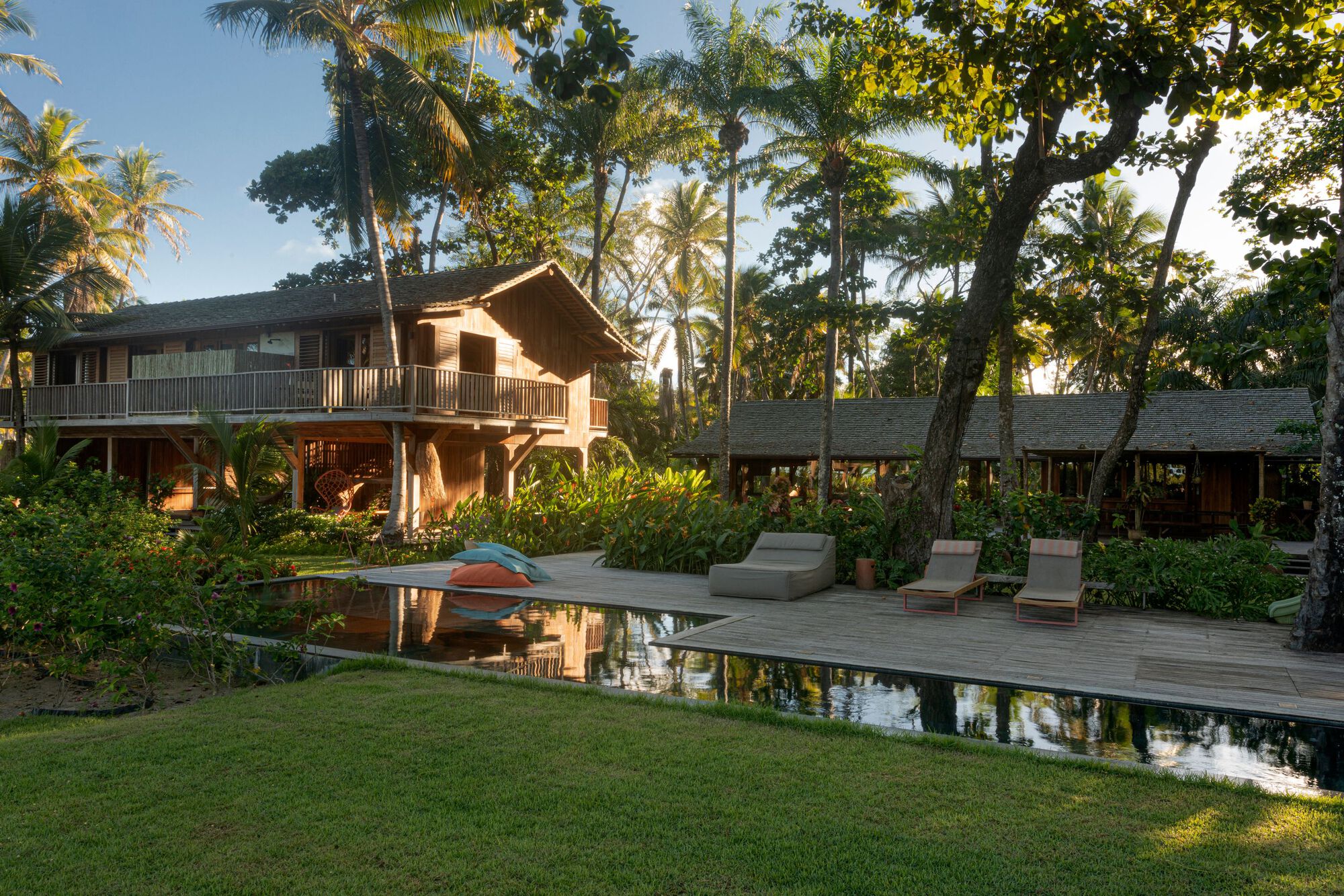 Wooden pool deck with lounge chairs facing the timber house and thatched pavilion, palms reflected in the still water