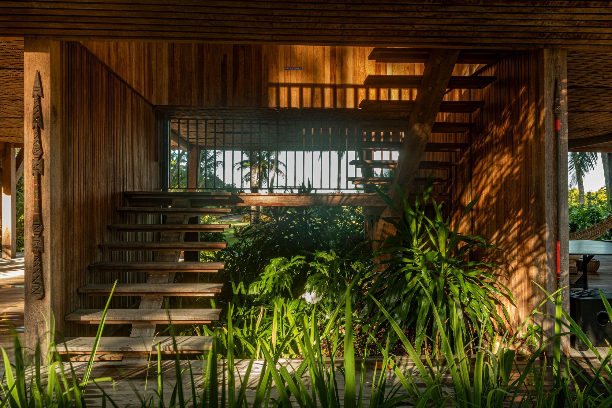 Open timber stair beneath the raised house, framed by carved post and tall grasses in dappled sunlight