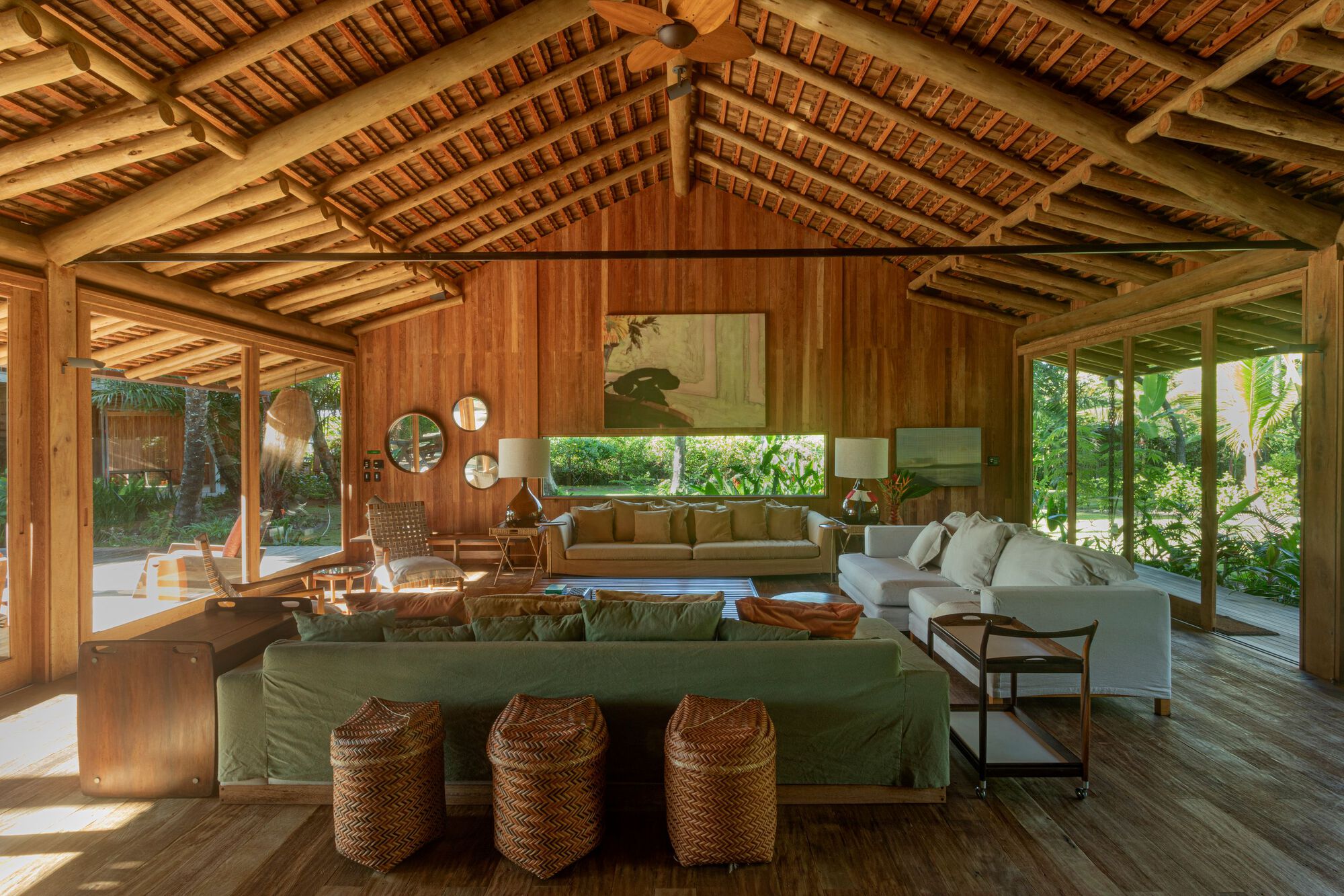 Living room under a round-log roof frame, with green sofa, woven stools and glazed walls opening to the garden