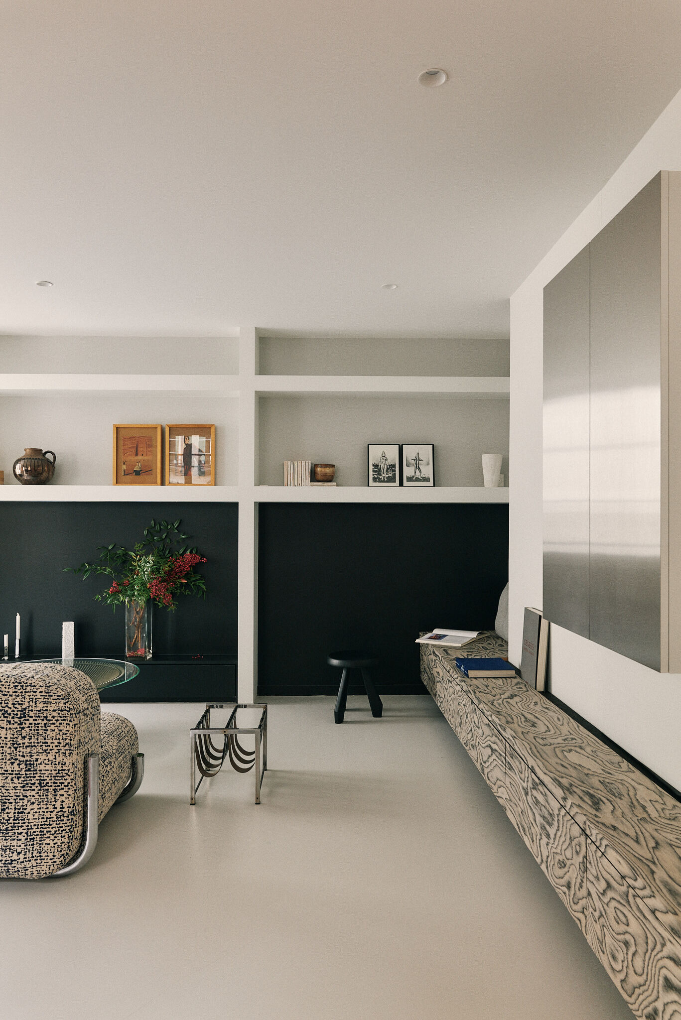 Black-backed white shelving with ceramics, framed prints and a red-berried vase beside a long floating wood bench and steel wall panel