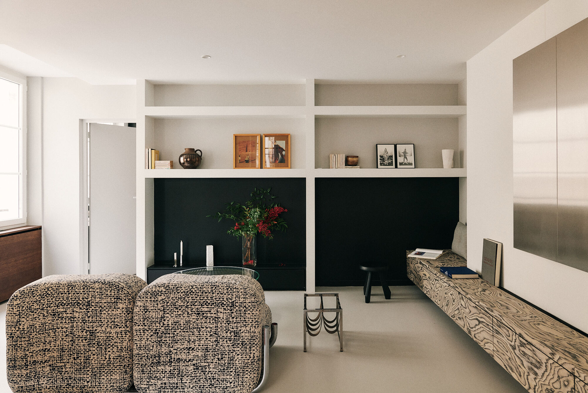 Two boucle ottomans face a white shelving unit with a black back panel, a stool and a floating swirl-grain bench in daylight