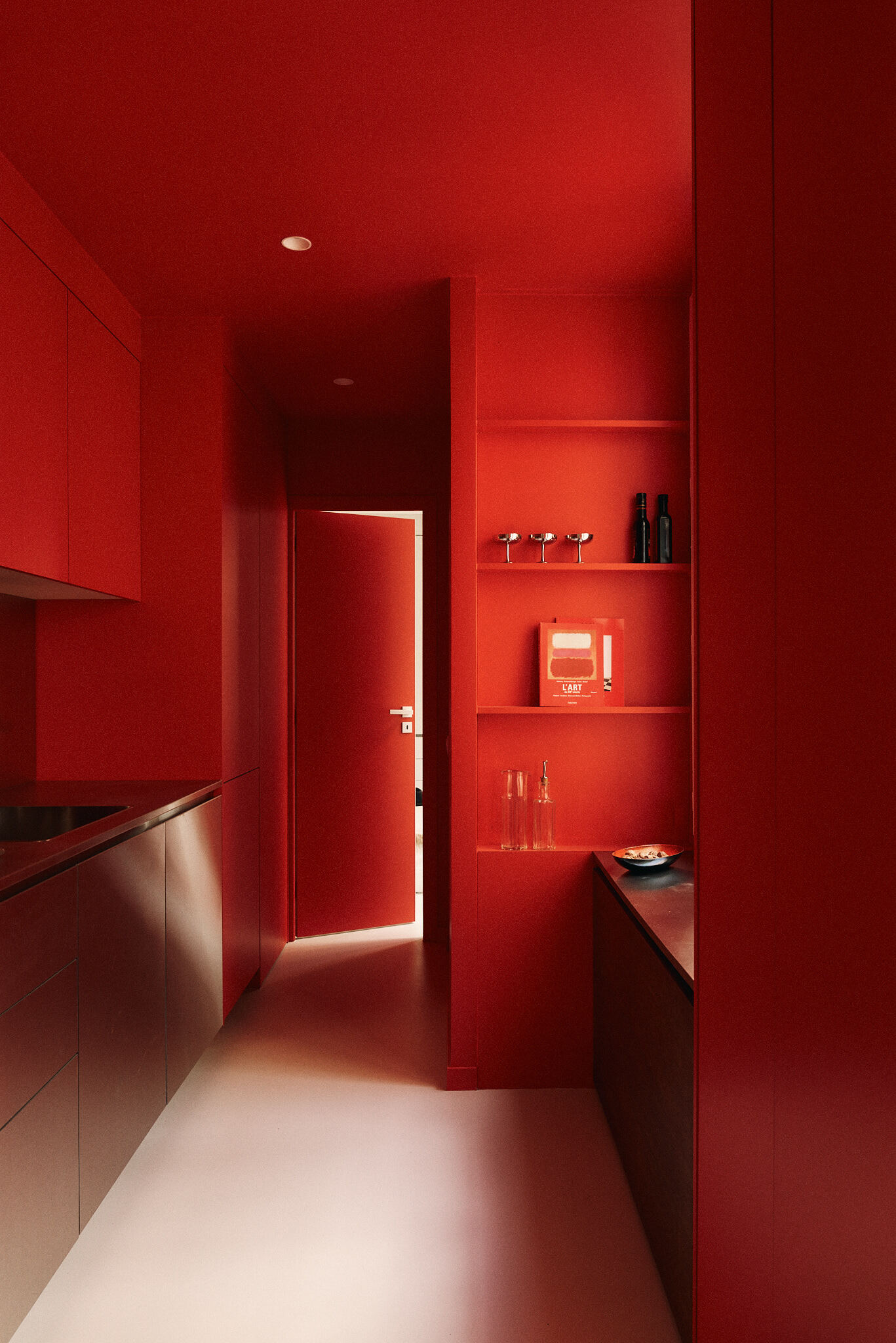 Narrow kitchen corridor painted entirely in saturated red, with steel base cabinets, open shelves and a red door glowing at the far end