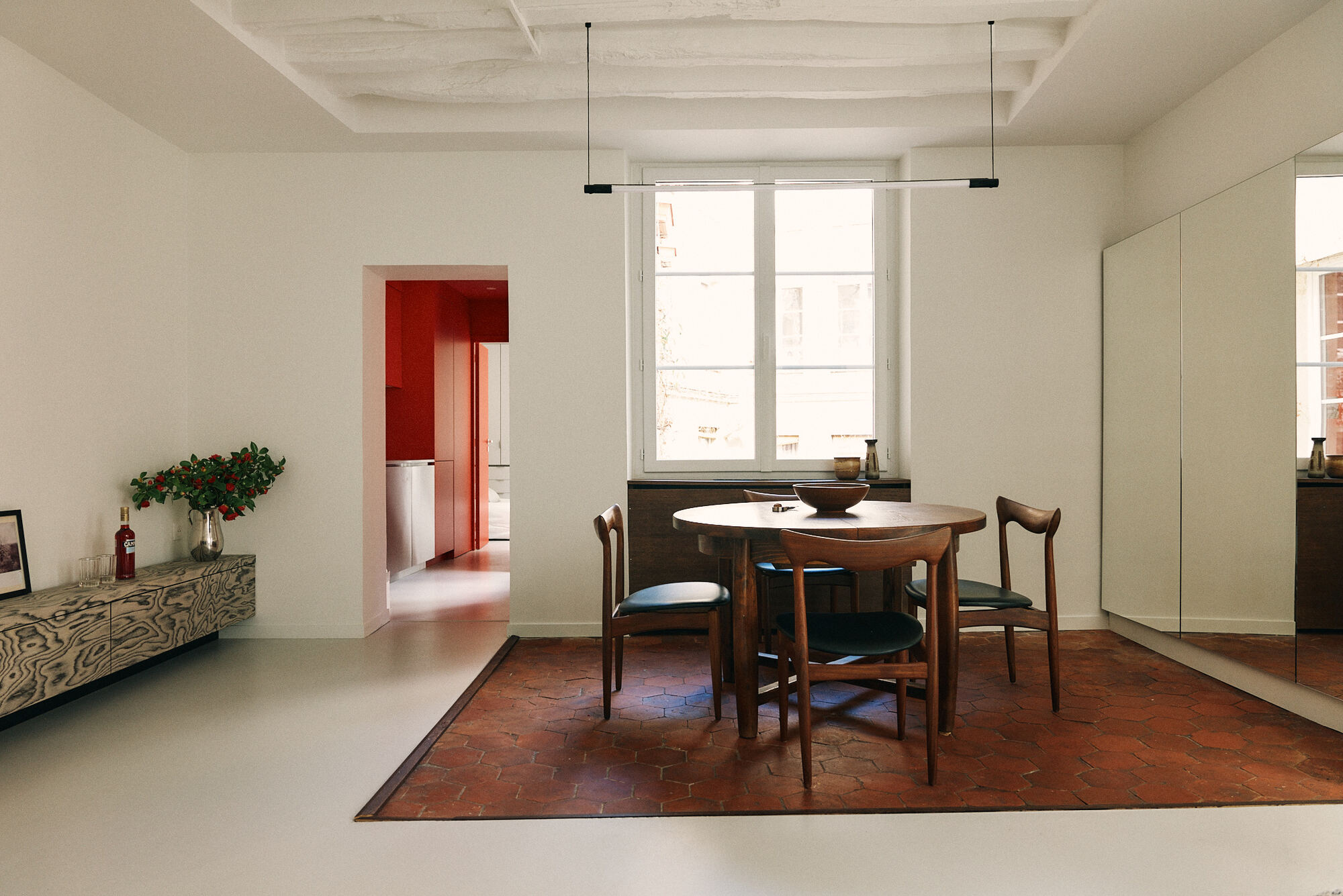 Round timber dining table and four chairs on a hexagonal terracotta tile inset, with a window behind and a red room visible through the doorway